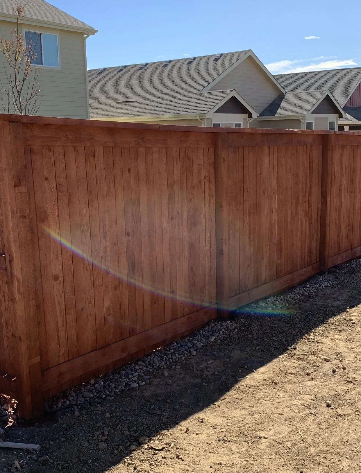 Newly installed wooden privacy fence in a backyard with neighboring houses visible in the background, under a clear blue sky.