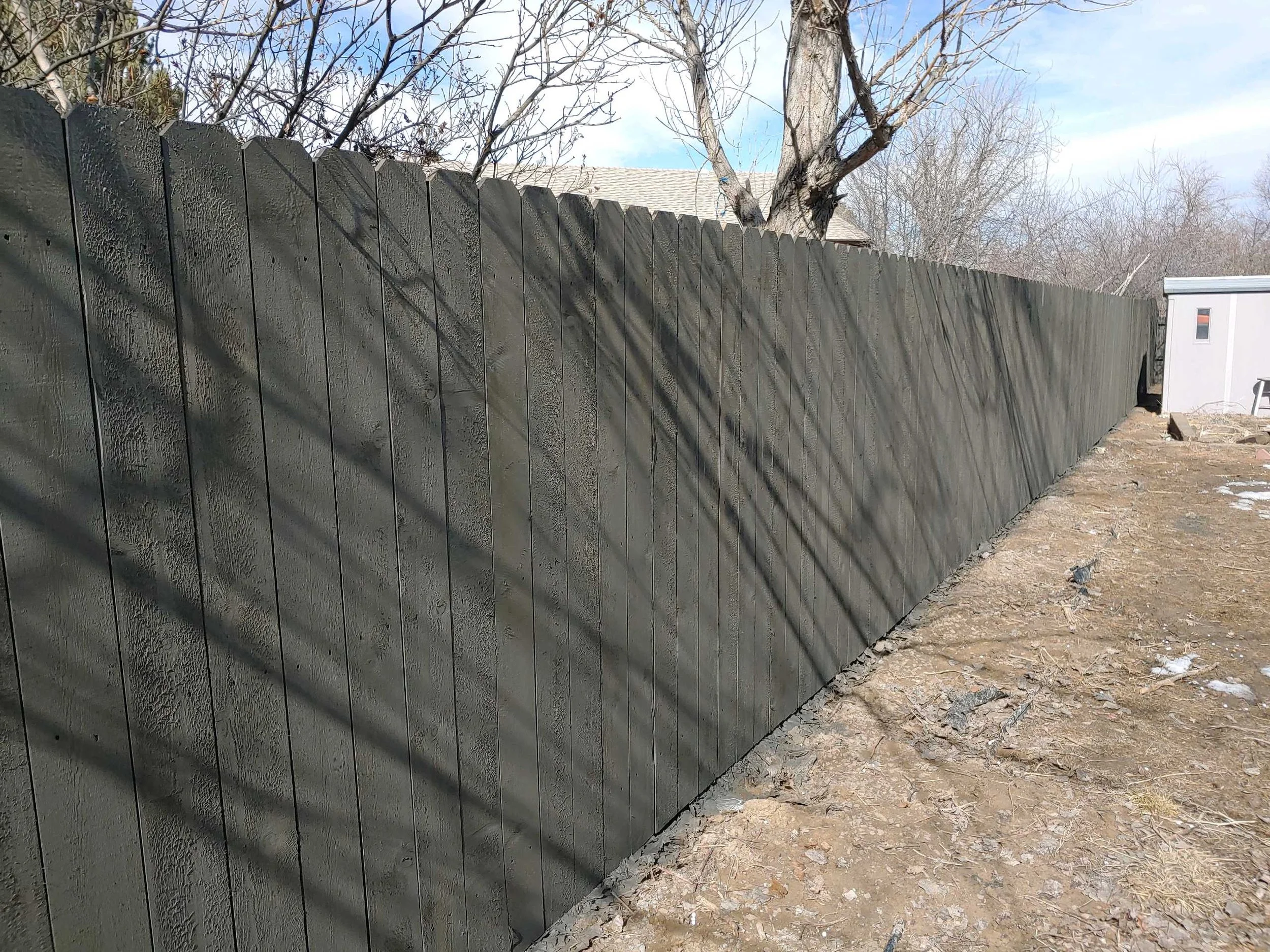 Outdoor scene with a new dark green wooden fence, tree with no leaves, and a small white shed in the background. Shadows of tree branches cast on the fence, dirt ground with some patches of snow.