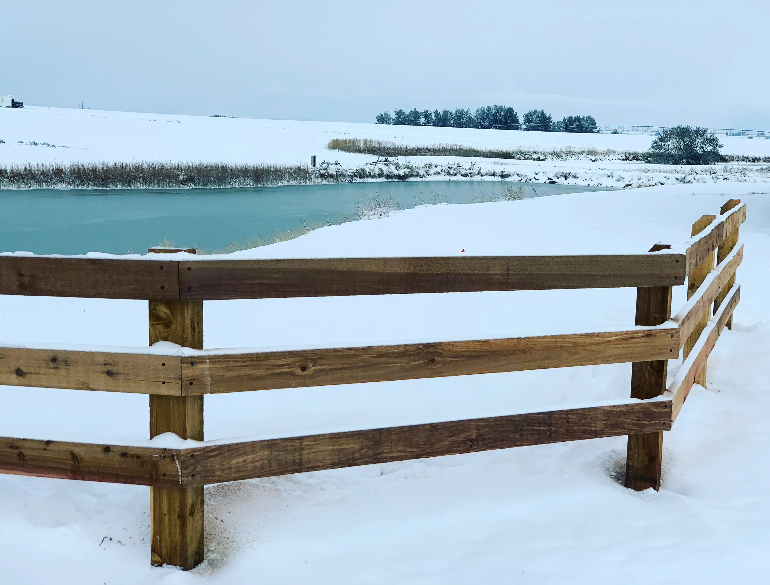 A snow-covered landscape with a wooden fence in the foreground, a partially frozen pond or small lake, and fields with snow and some trees in the background.