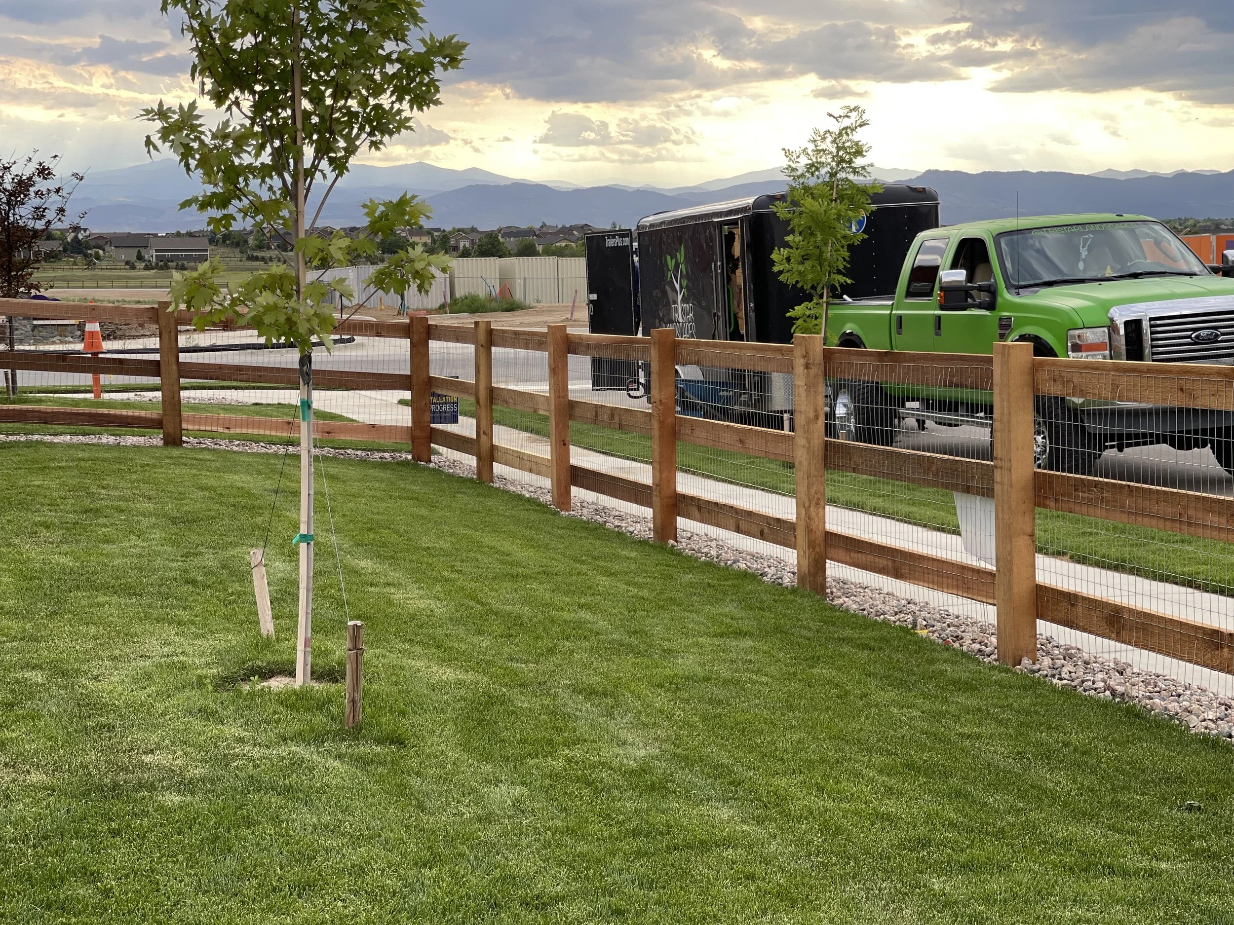A grassy yard with newly planted trees, wooden fencing, a sidewalk, and a green truck parked beside a black trailer in the background, with mountains and a cloudy sky in the distance.