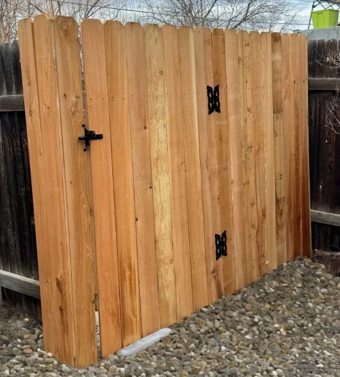 Newly installed wooden fence with black metal hinges and latch, placed on gravel ground in backyard.
