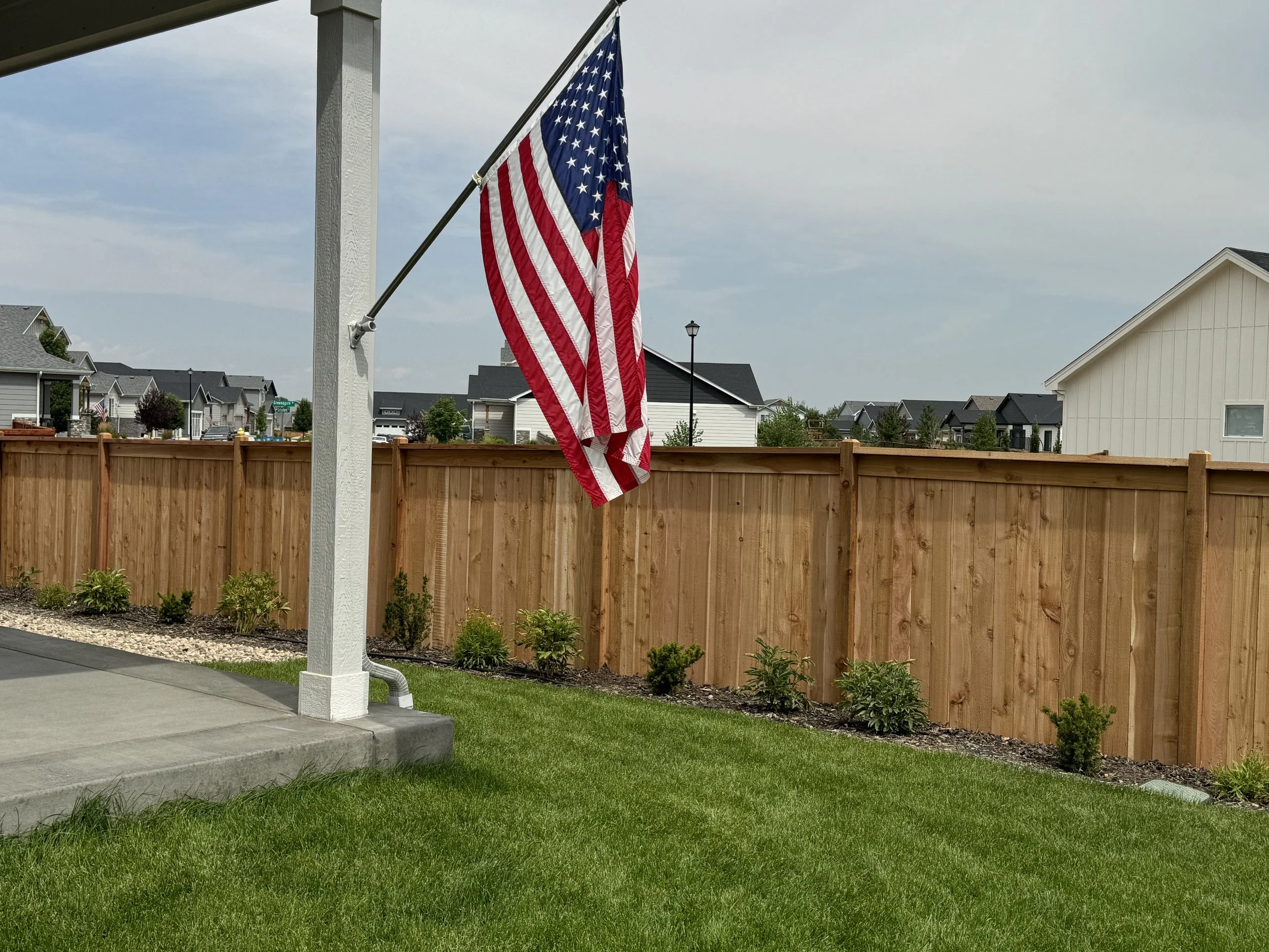 American flag hanging on a flagpole in a backyard with a wooden fence, green lawn, and neighboring houses in the background.