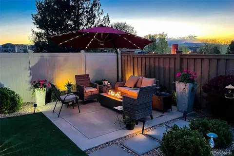 Backyard patio with outdoor furniture, a fire pit, umbrella, potted plants, and a grassy area, during sunset.