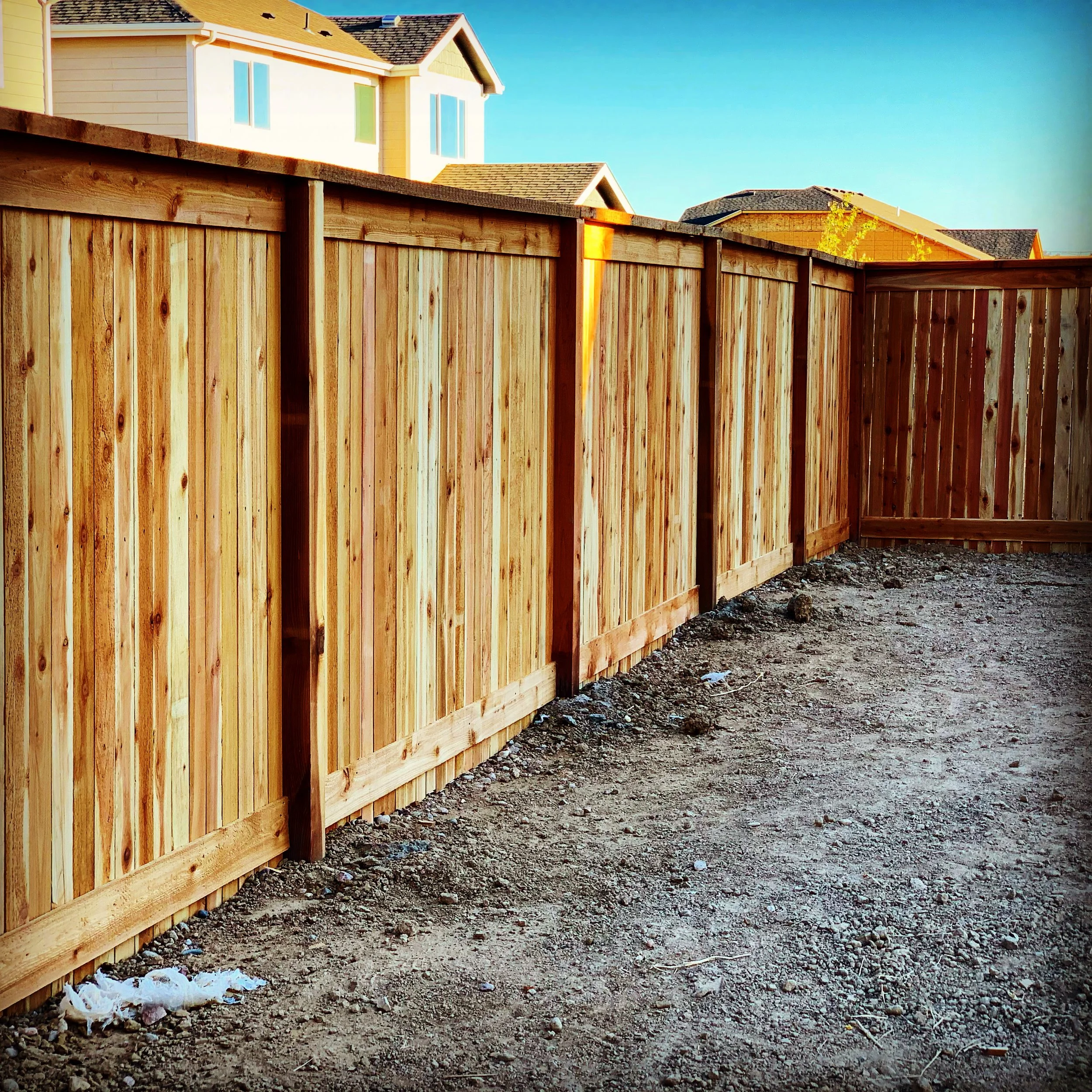 A newly built wooden privacy fence in a backyard, with houses and a clear blue sky in the background.