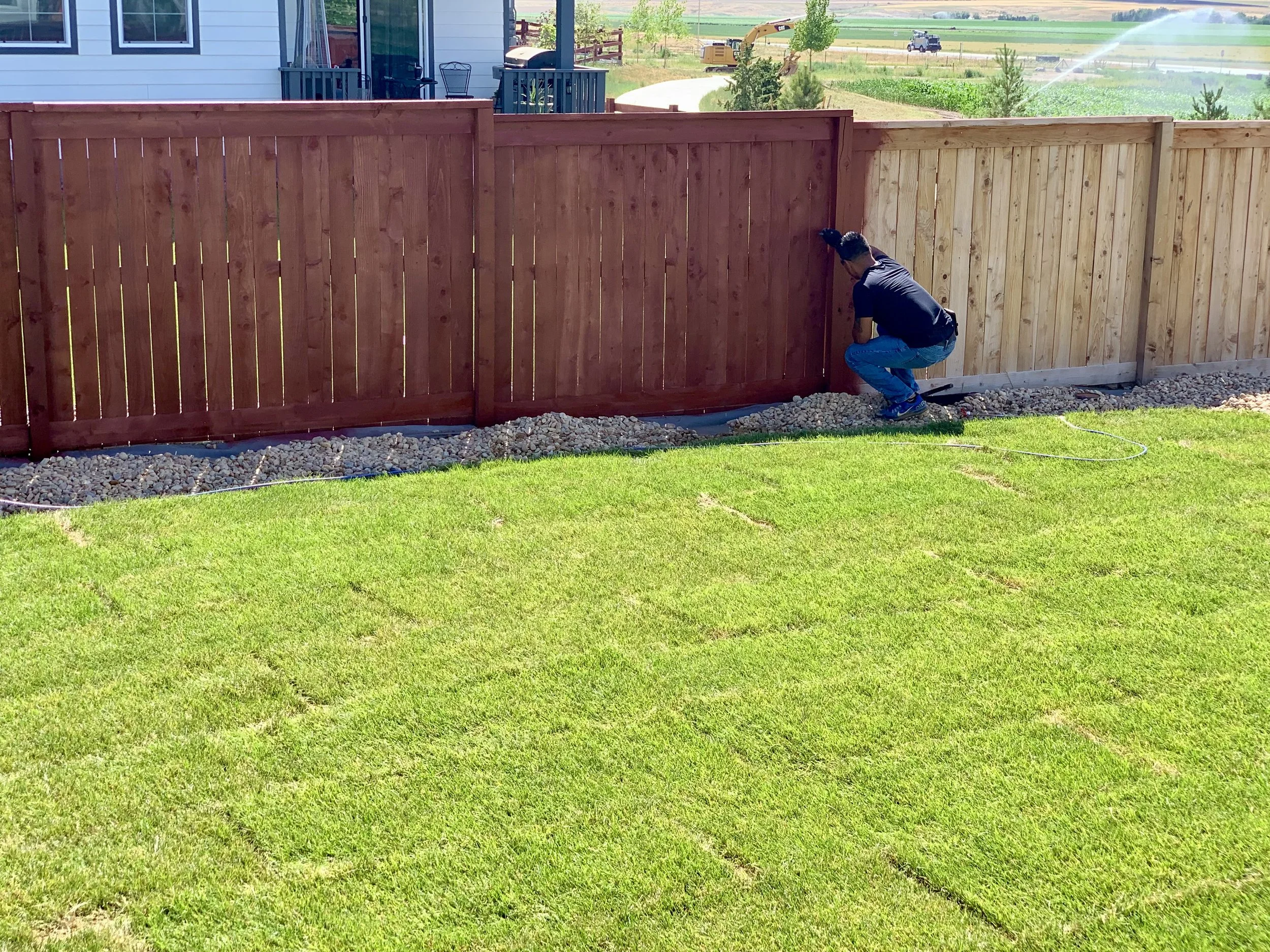 Person installing a wooden fence in a backyard, with a green lawn and gravel base, while sunlight illuminates the area.