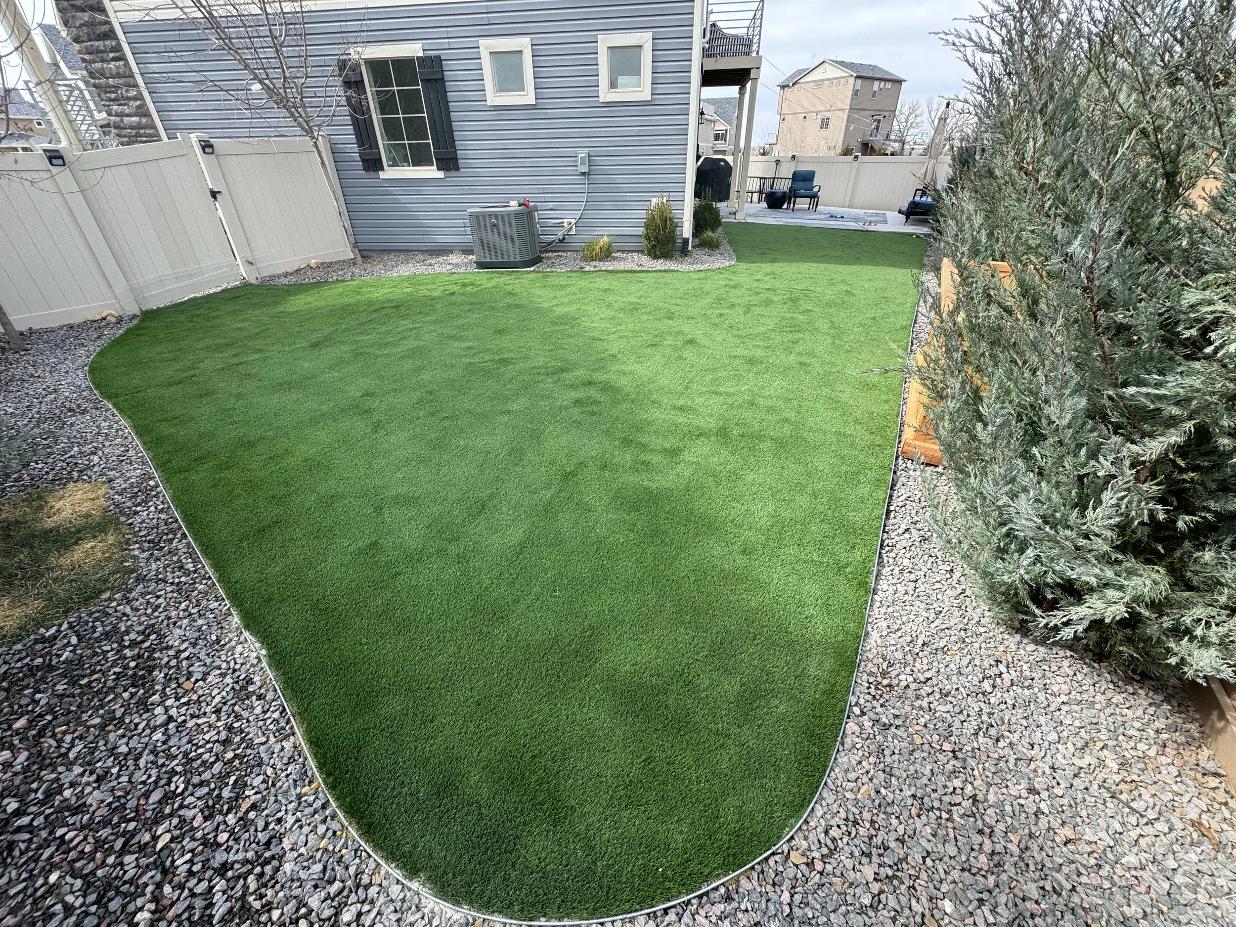 Backyard with artificial grass lawn, landscaped with gravel borders, a beige fence, and a gray house with black shutters, outdoor seating area, and trees.