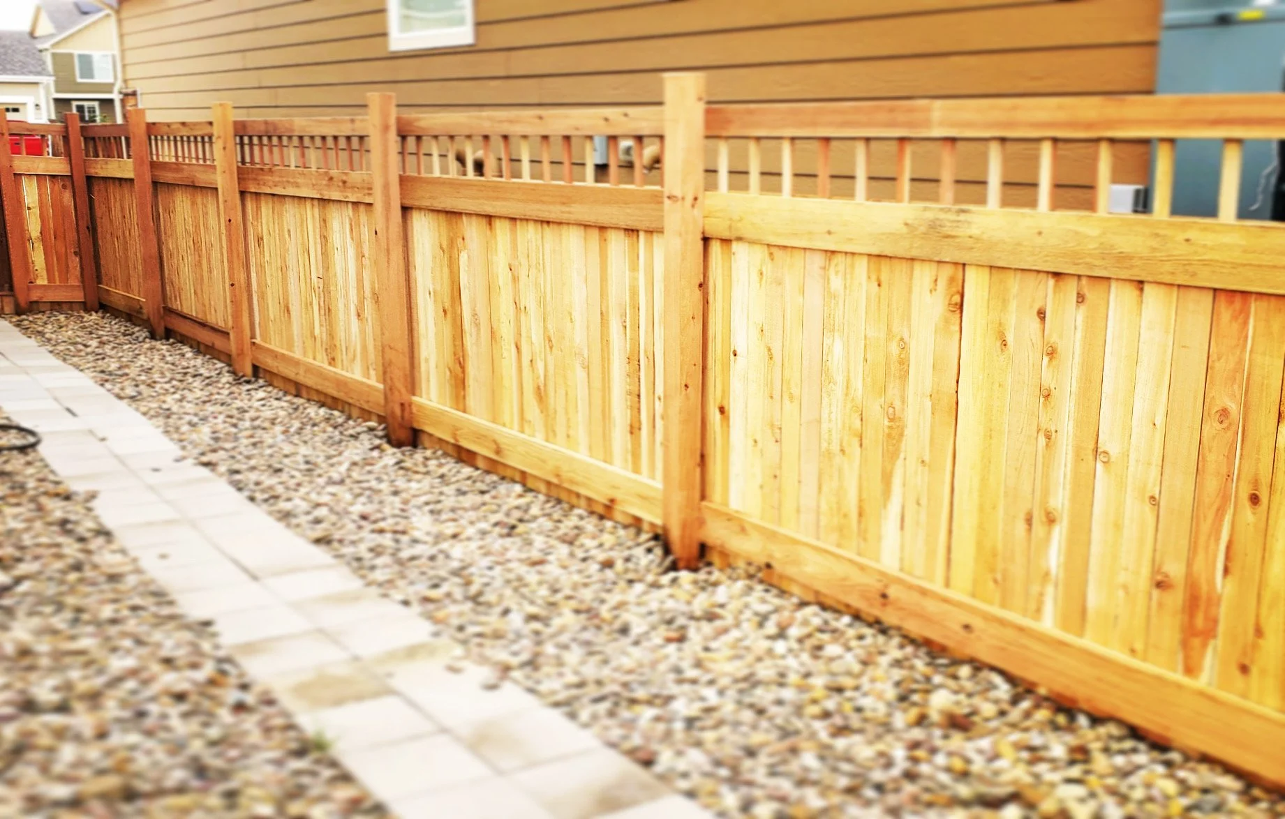 Wooden privacy fence alongside a pathway with gravel and pavers, outside residential buildings.