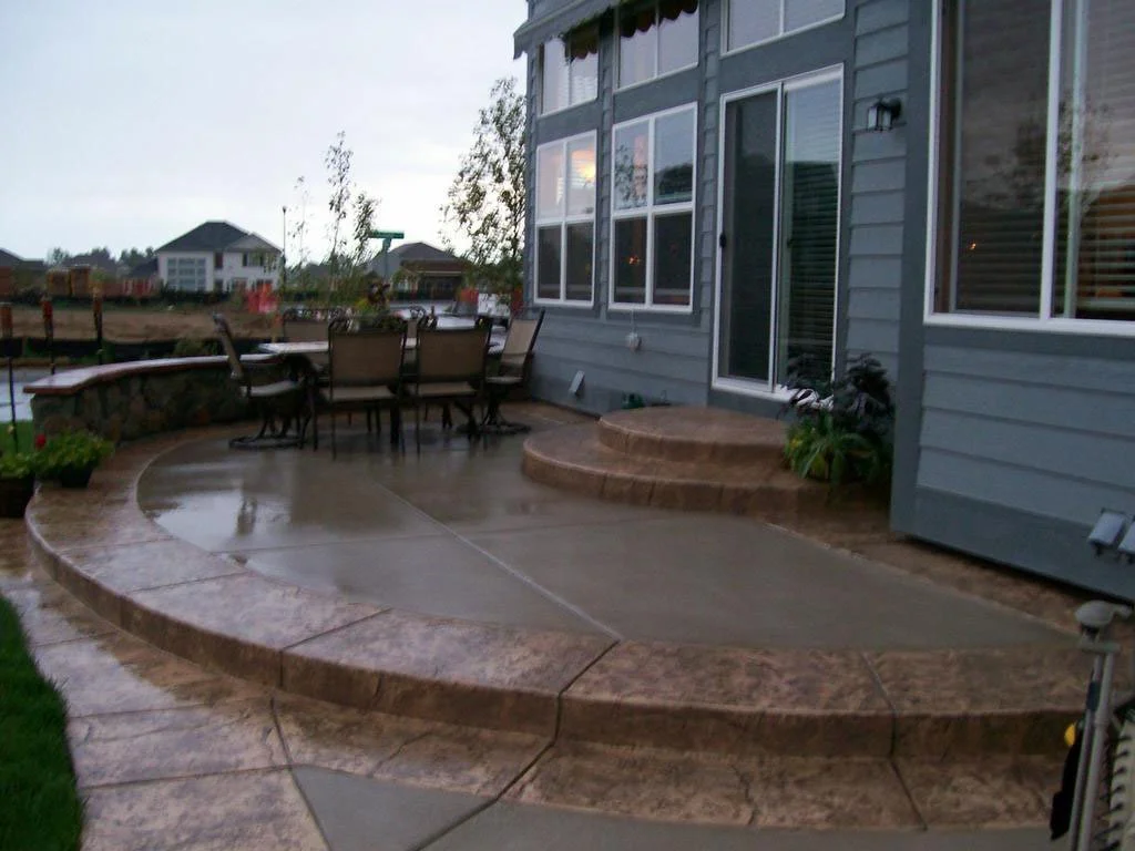 Outdoor concrete patio with stone steps leading to a sliding glass door, a patio table with chairs, and neighboring houses visible in the background.