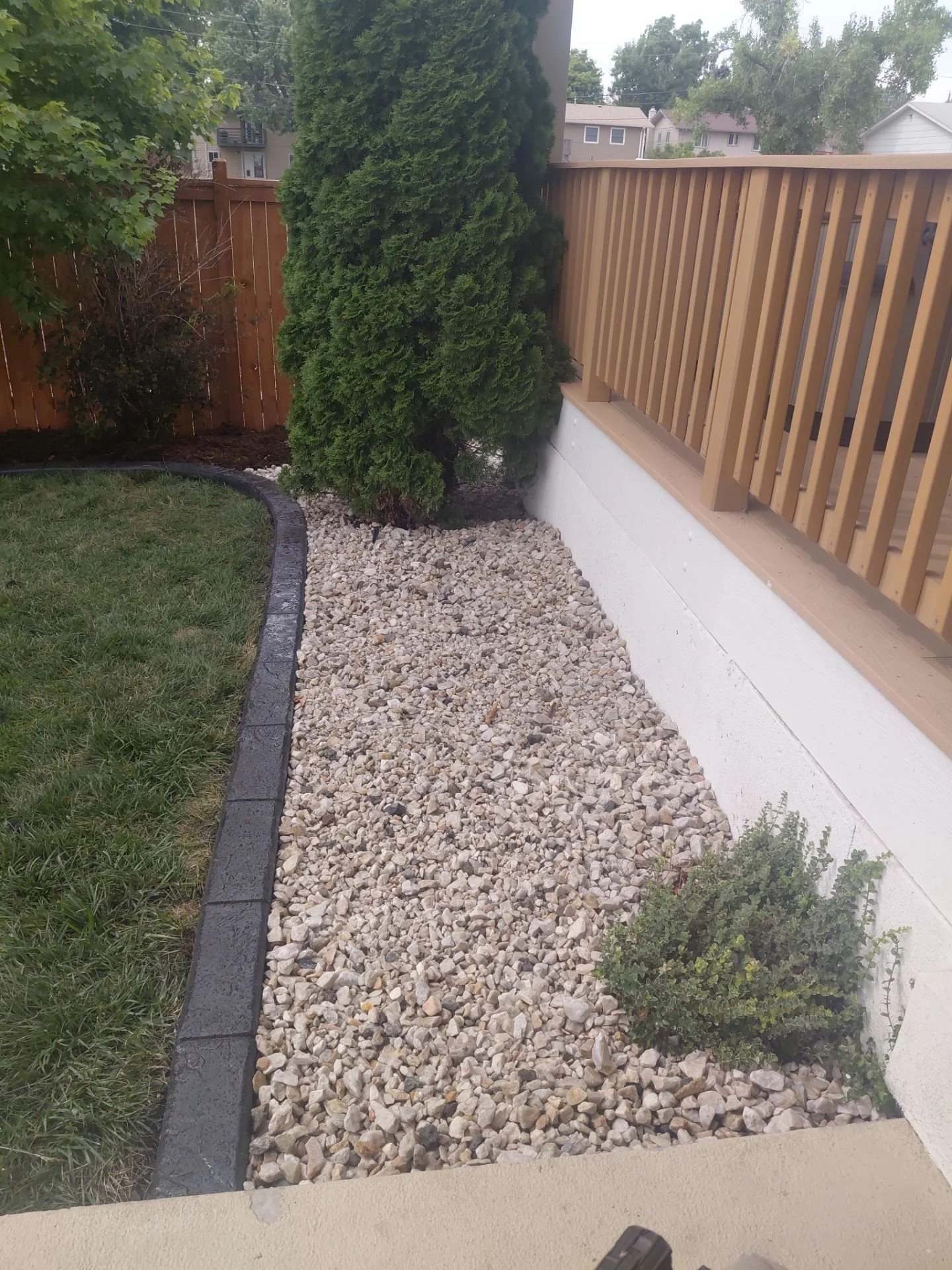 A backyard corner with grass, a tree, a rock border, and small bushes, fenced with a wooden railing and a privacy fence.