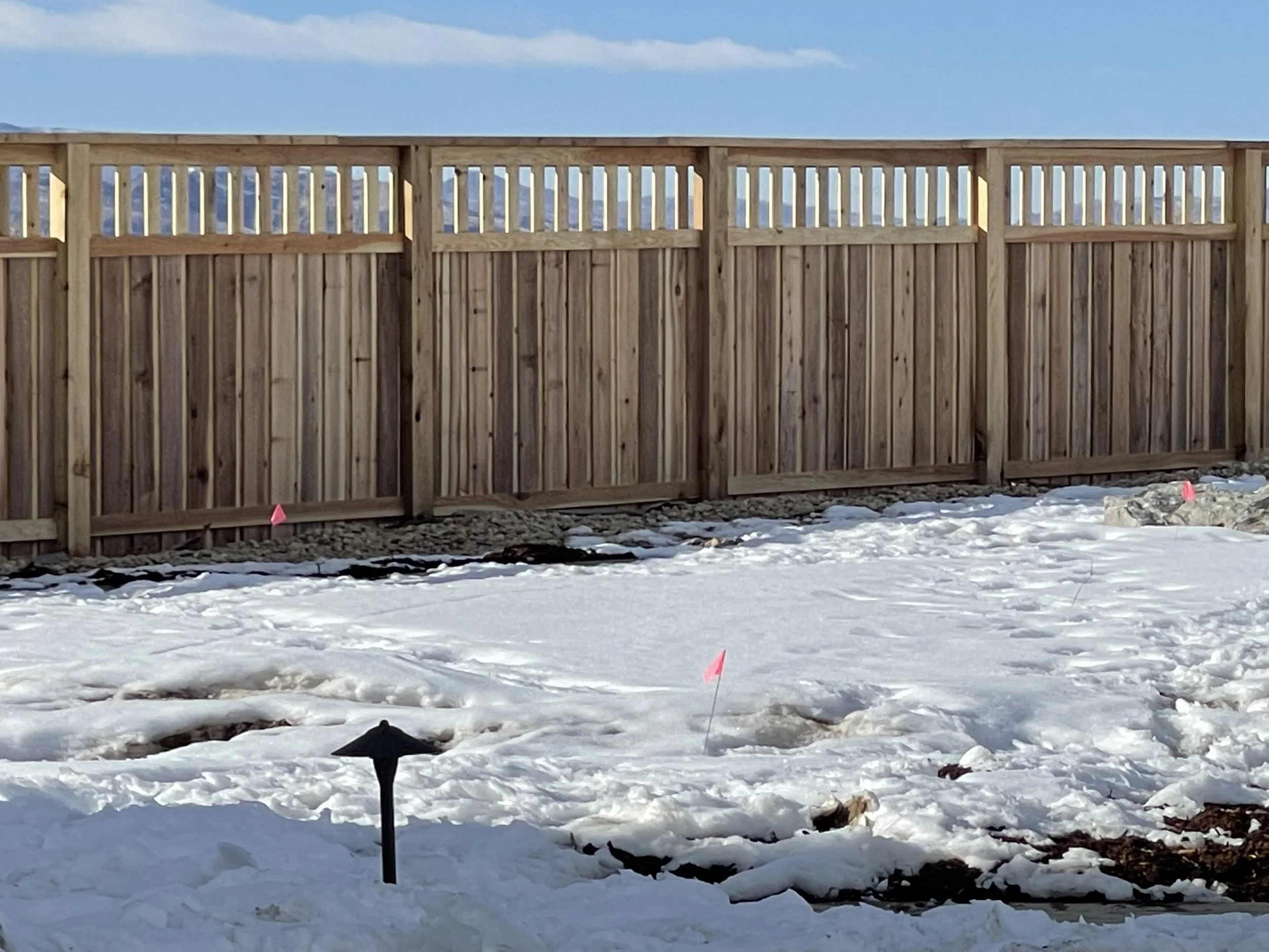 New custom HOA standard wooden fence on a snowy landscape with pink marking flags, black landscape light, and a blue sky.