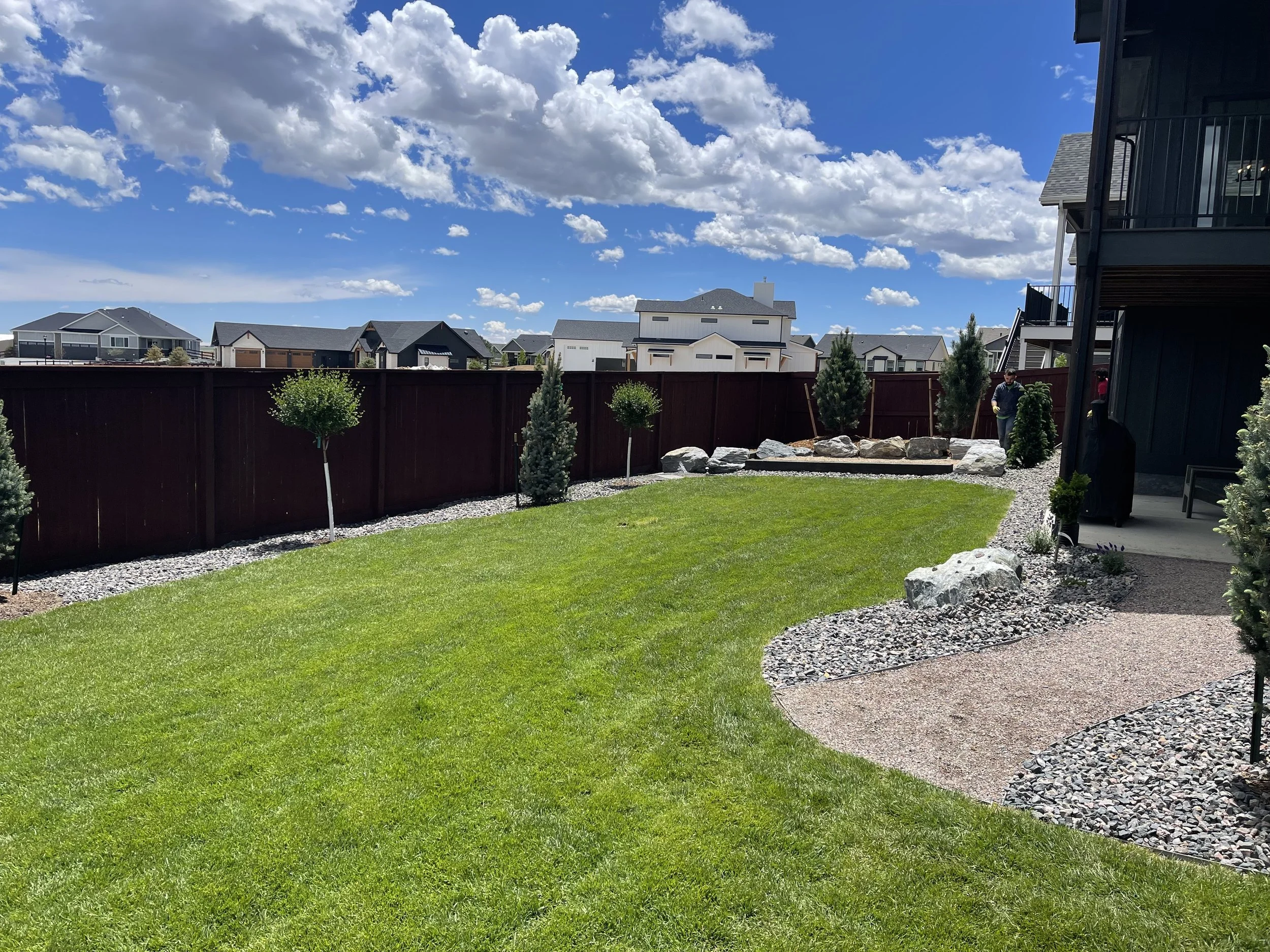 A backyard with green grass, small trees, rocks, and a red fence. On the right, part of a house with a balcony and a person standing near the trees. The sky is blue with scattered white clouds.
