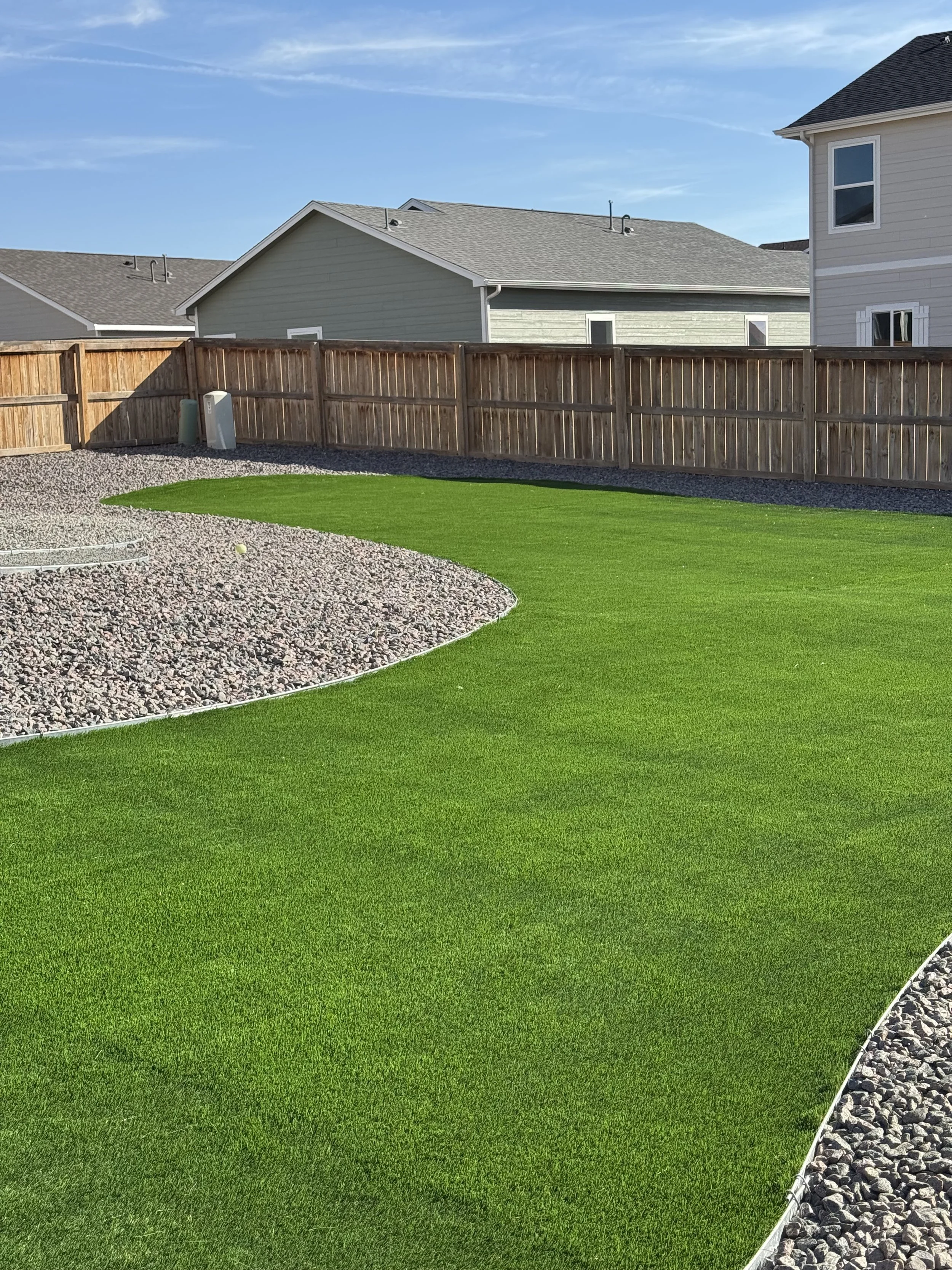 A backyard with a well-maintained green lawn, a gravel area, a wooden fence, and neighboring houses under a blue sky.