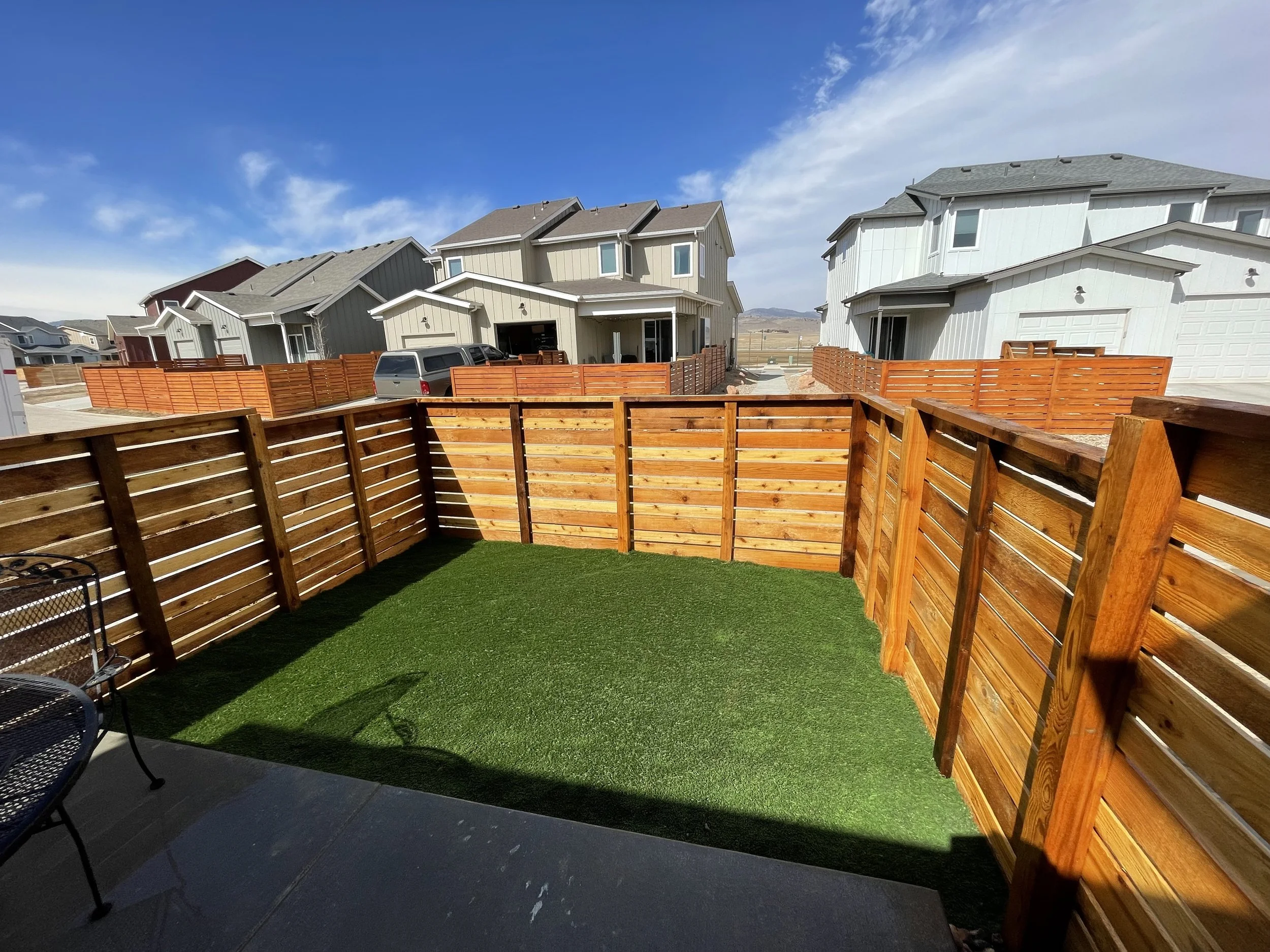 Backyard with artificial grass, wooden fence, patio furniture, and neighboring houses under a blue sky.