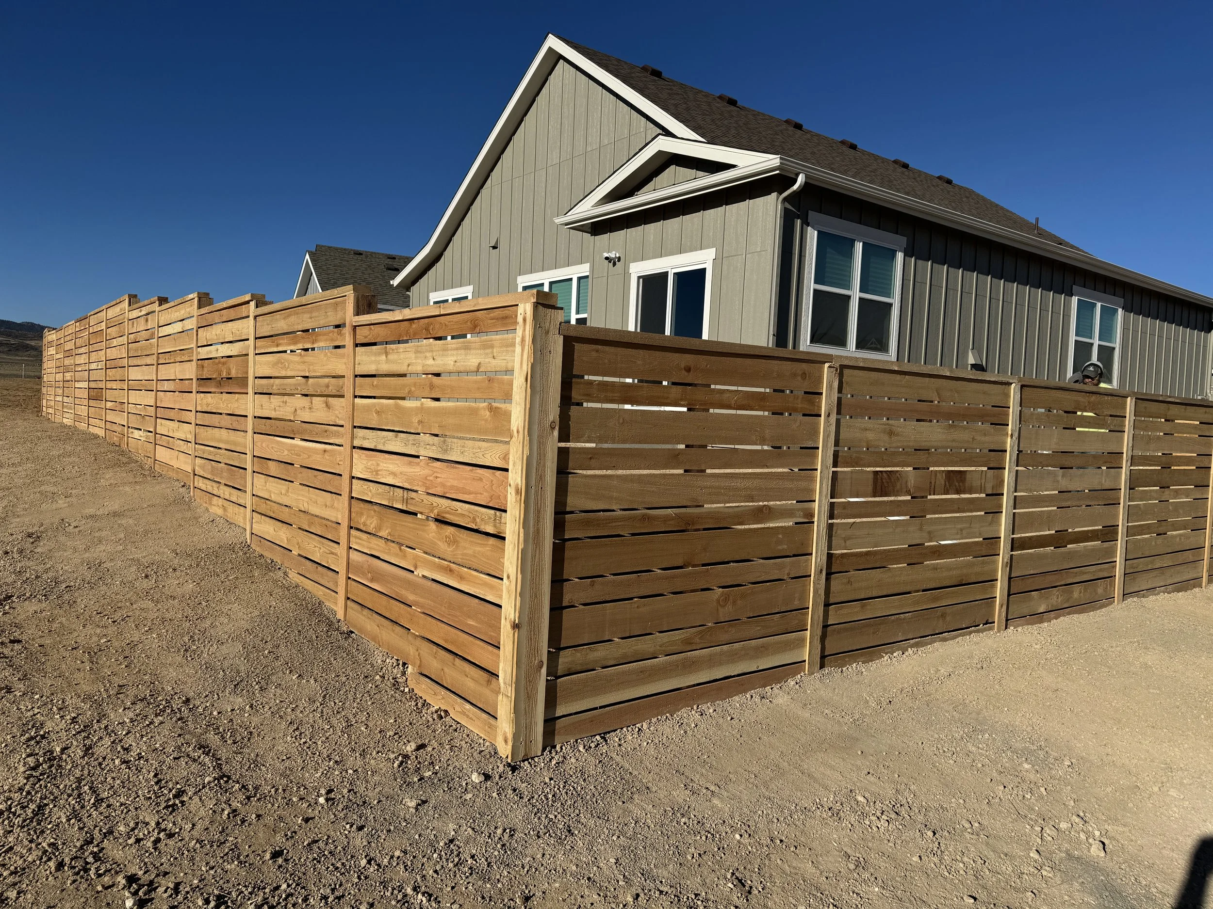 New wooden fence beside a gray house with multiple windows, under a clear blue sky.