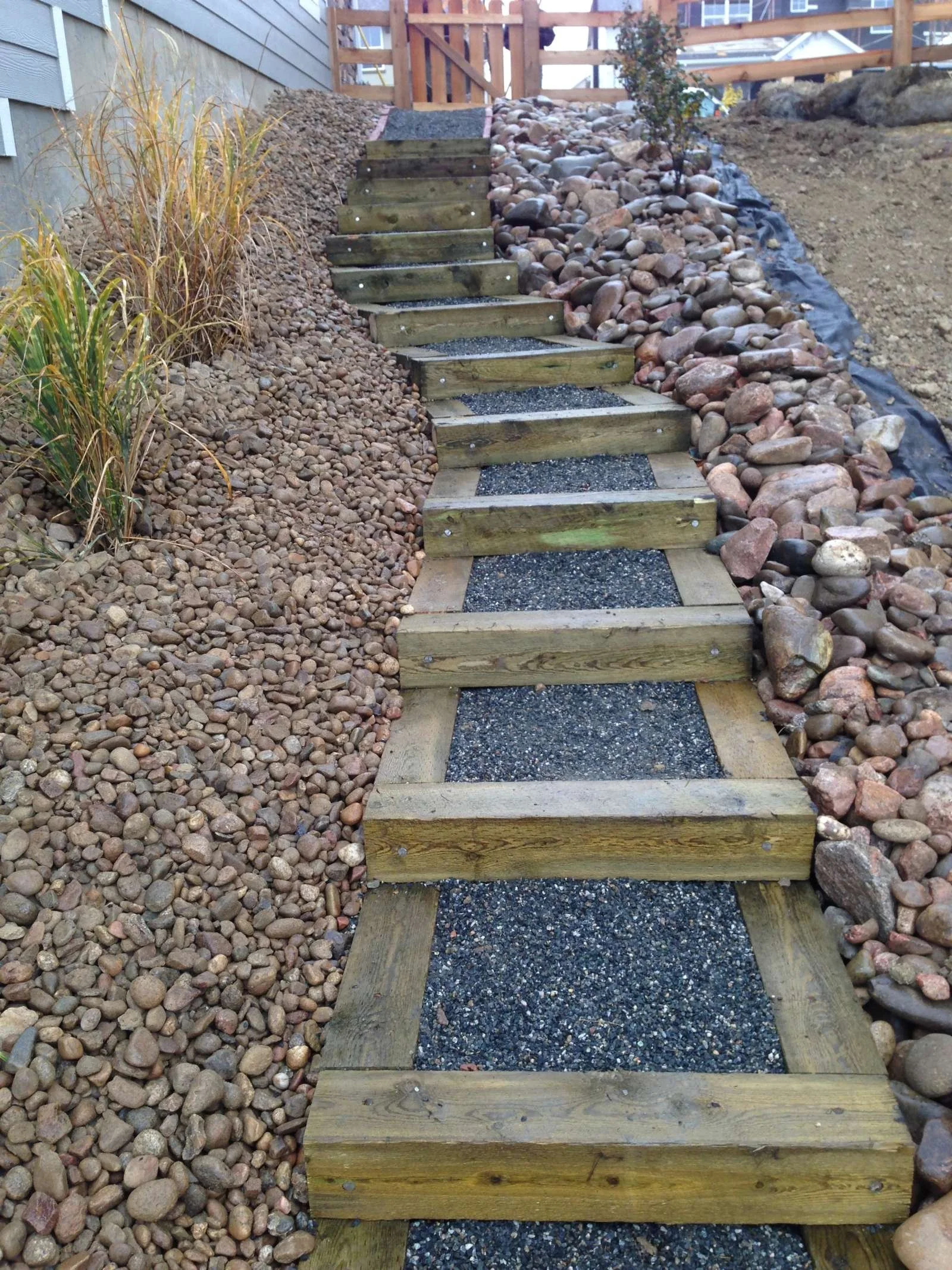 A set of wooden steps with sand and gravel filling the spaces between the treads, leading uphill. The surrounding landscaping includes small pebbles, mulch, and some ornamental grasses on the left, with larger rocks on the right. A wooden fence is at