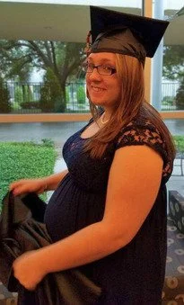 Pregnant woman wearing a graduation cap and gown, smiling slightly as she holds part of her gown. She is dressed in a dark lace dress, and stands indoors by a window with greenery visible outside.
