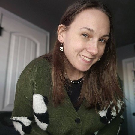 A young woman with brown hair and earrings smiling at the camera inside a house.