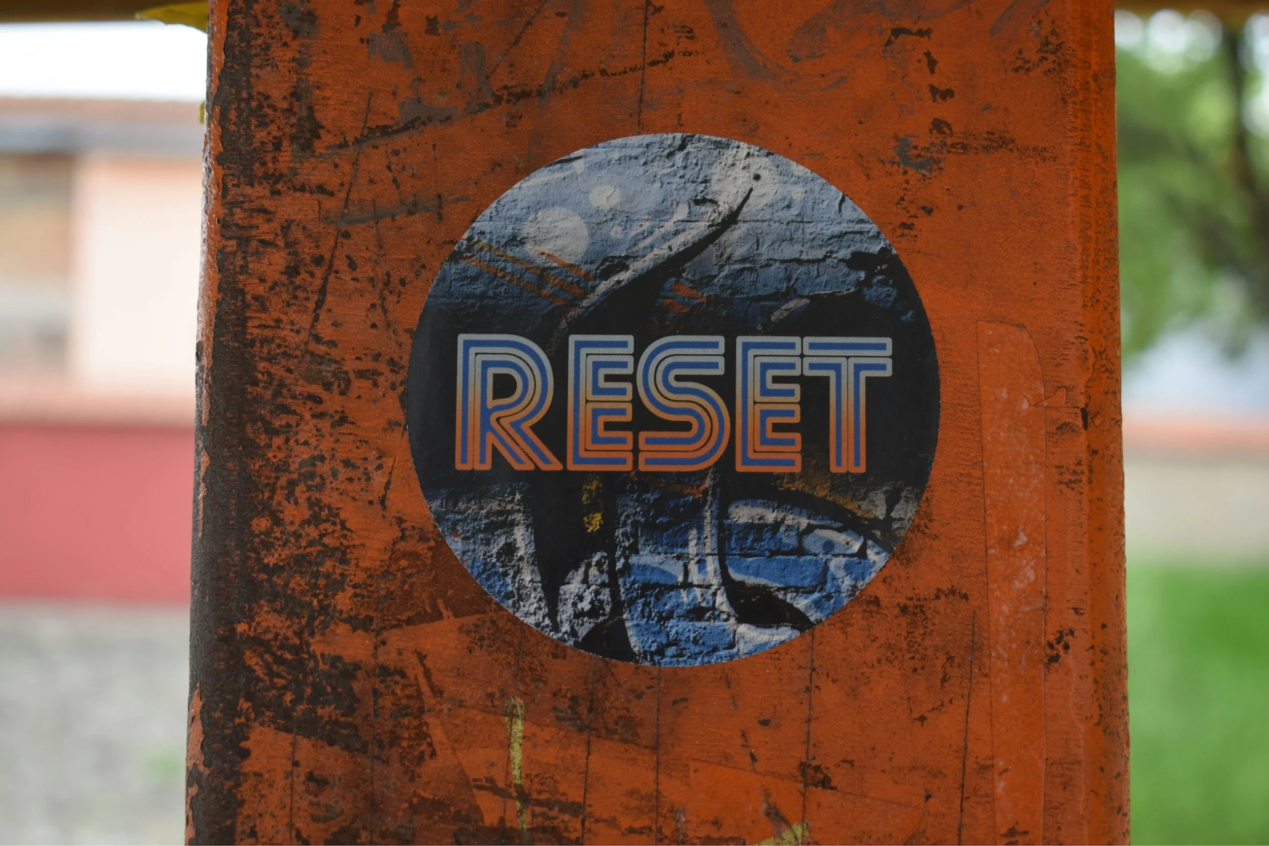 A round sticker with the word 'RESET' in bold, colorful text, on a rusty orange metal surface with some scratches and peeling paint.