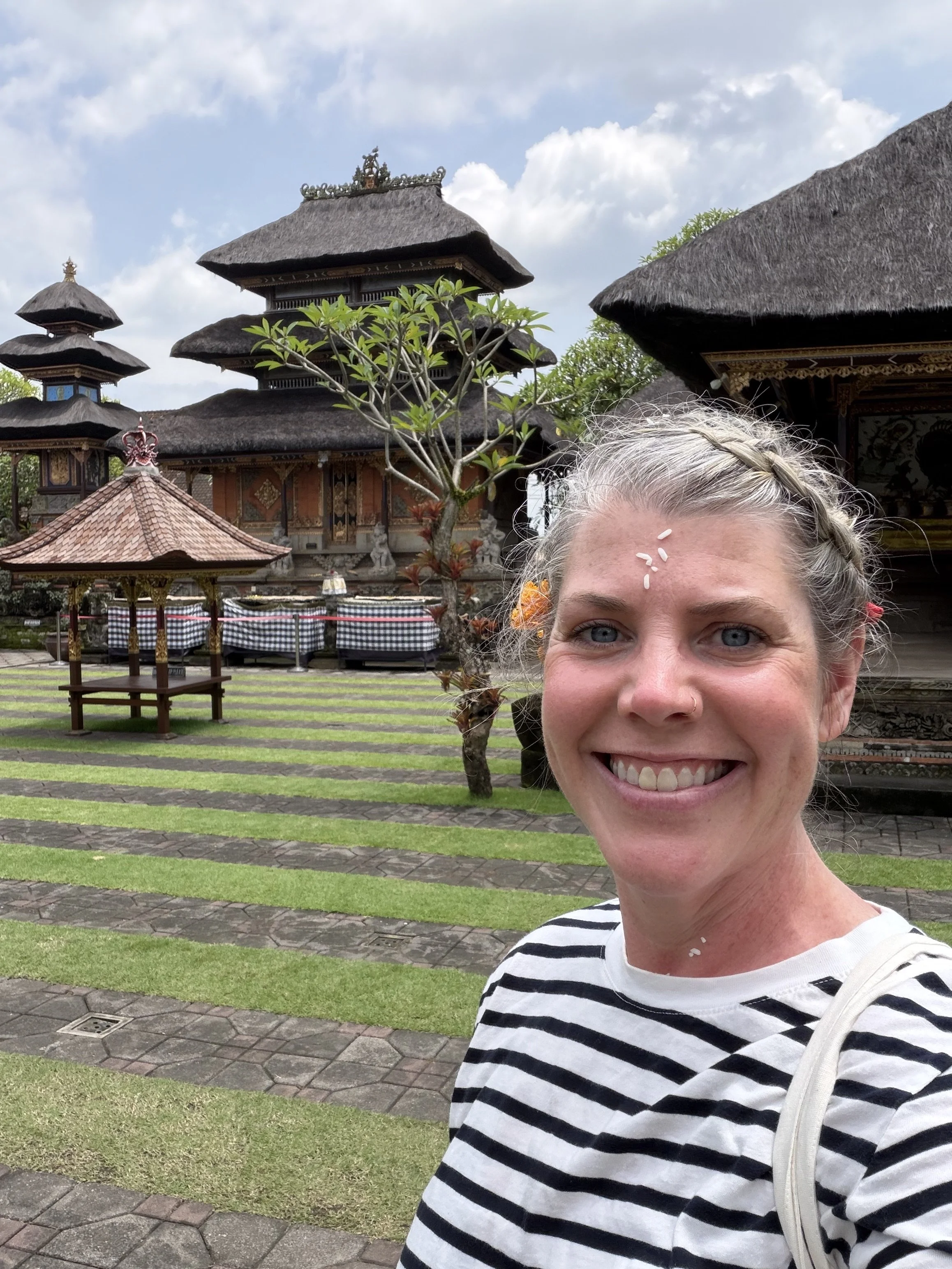 A woman with blonde hair in a braid, wearing a striped shirt, smiling while taking a selfie outside in front of traditional Balinese architecture with thatched roofs and a tree, under a partly cloudy sky.