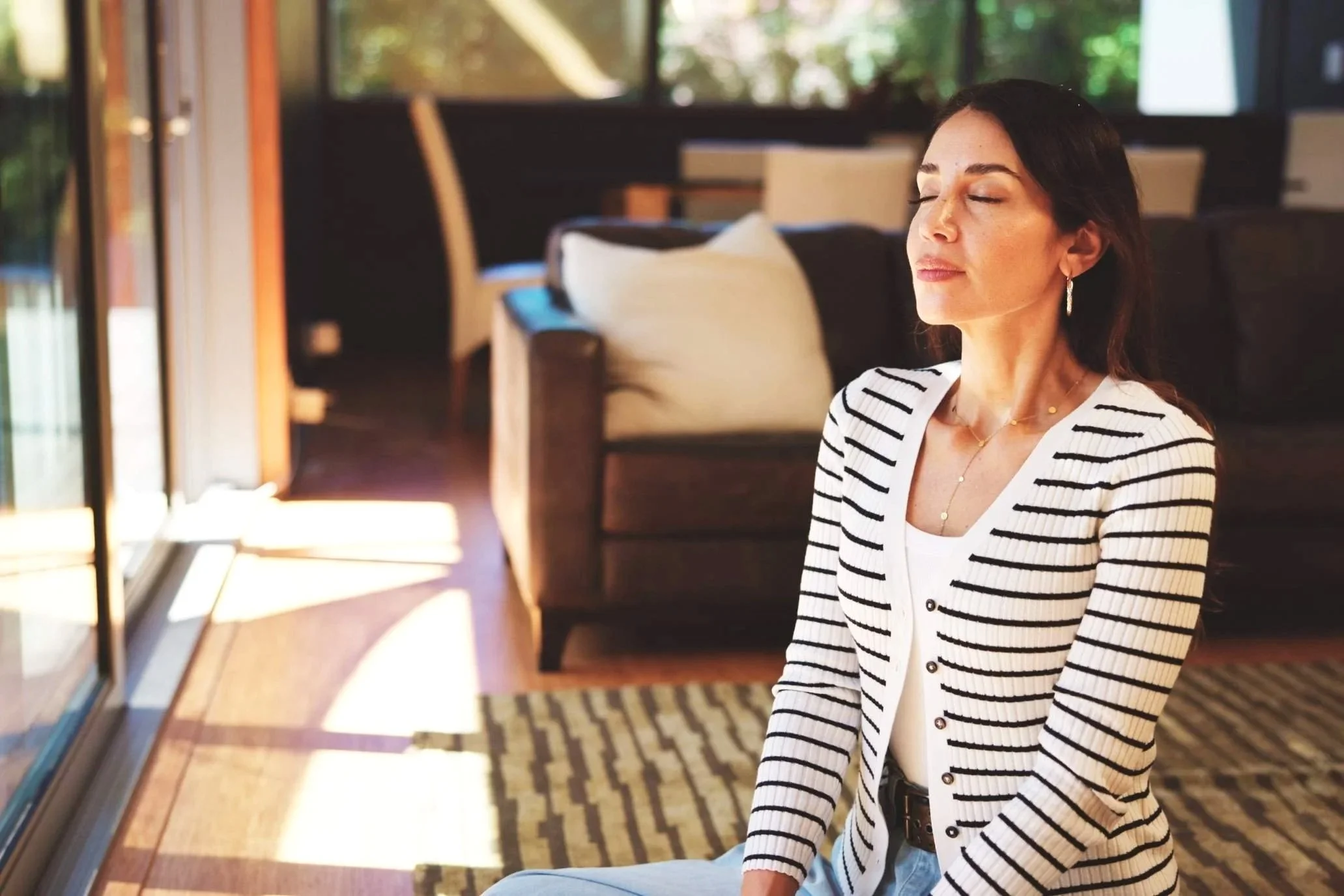 A woman with dark hair and earrings, wearing a striped blazer, sitting with her eyes closed in a room with large windows and sunlight streaming in.