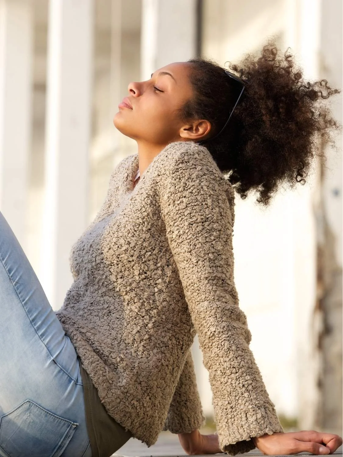 A woman with curly hair, wearing sunglasses on her head and a beige fuzzy sweater, is sitting outdoors with her eyes closed, leaning back and enjoying the sunlight.
