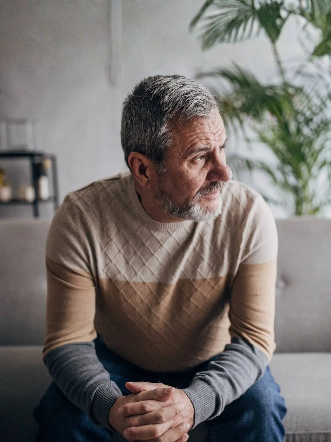 An older man with gray hair and beard sitting indoors, gazing thoughtfully out of frame. He is wearing a beige sweater and jeans, with a modern, minimalistic background including a gray couch and green plants.