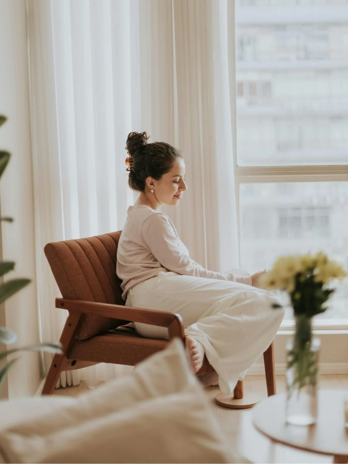 A woman with dark hair tied in a bun sitting on a brown armchair by a window with sheer white curtains, smiling with eyes closed and holding her legs, in a bright room with beige walls and a vase of flowers in the foreground.