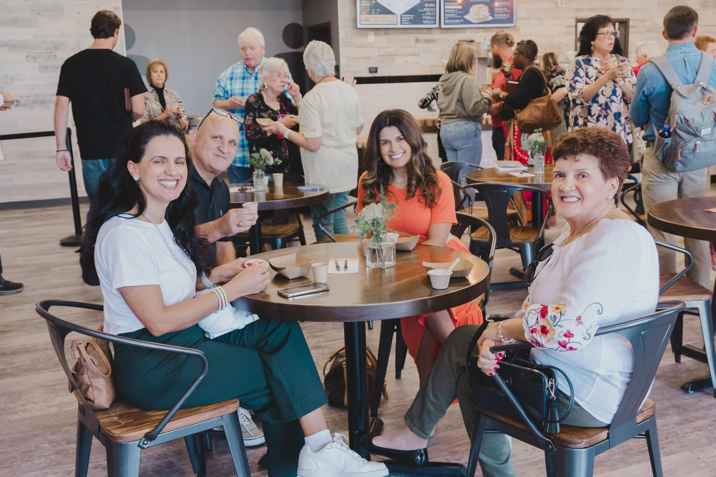 Four people seated at a round table in a busy cafe, smiling and enjoying their time together. The table has a small flower arrangement and some bowls with food or condiments. In the background, more people are standing and chatting in the cafe.