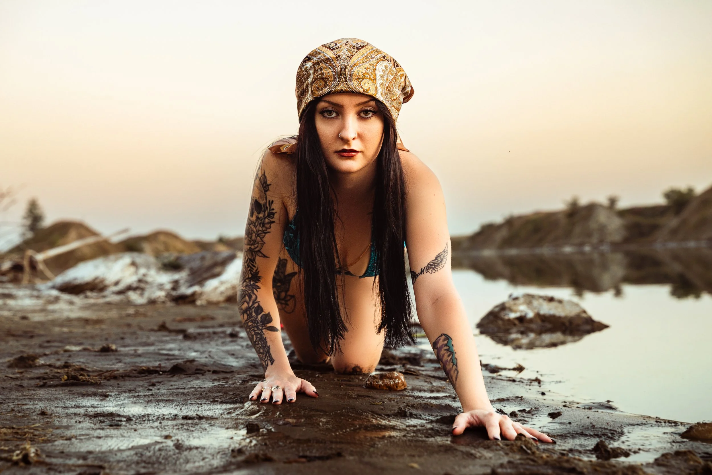 A woman with long dark hair, tattoos on her arms, and a patterned headscarf, kneeling on a rocky shoreline with water and distant hills at sunset near Calgary boudoir photography studio.