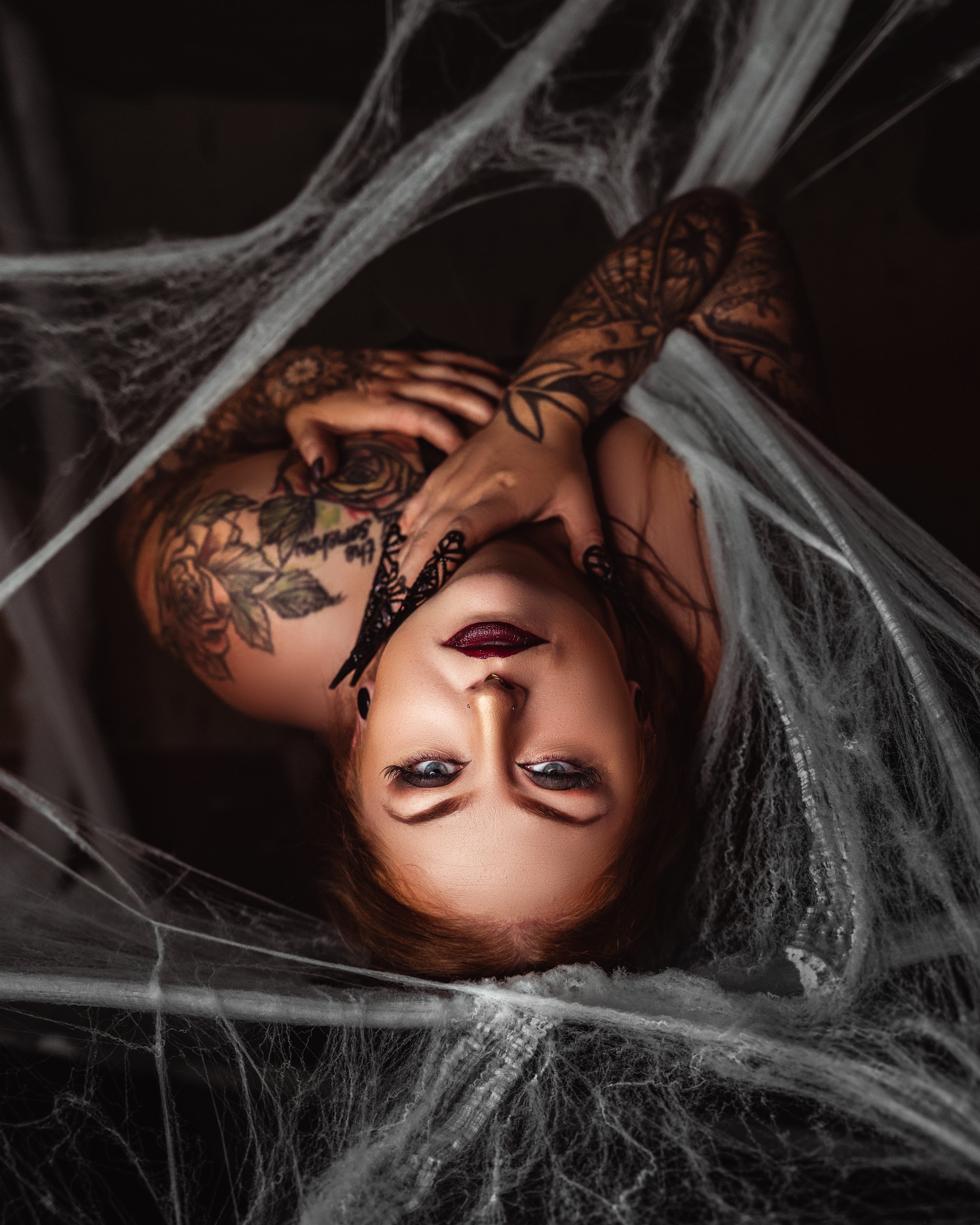 Woman lying upside down in a web of spider webs, with tattoos and dark makeup, looking at the camera in Calgary Boudoir Photography studio.