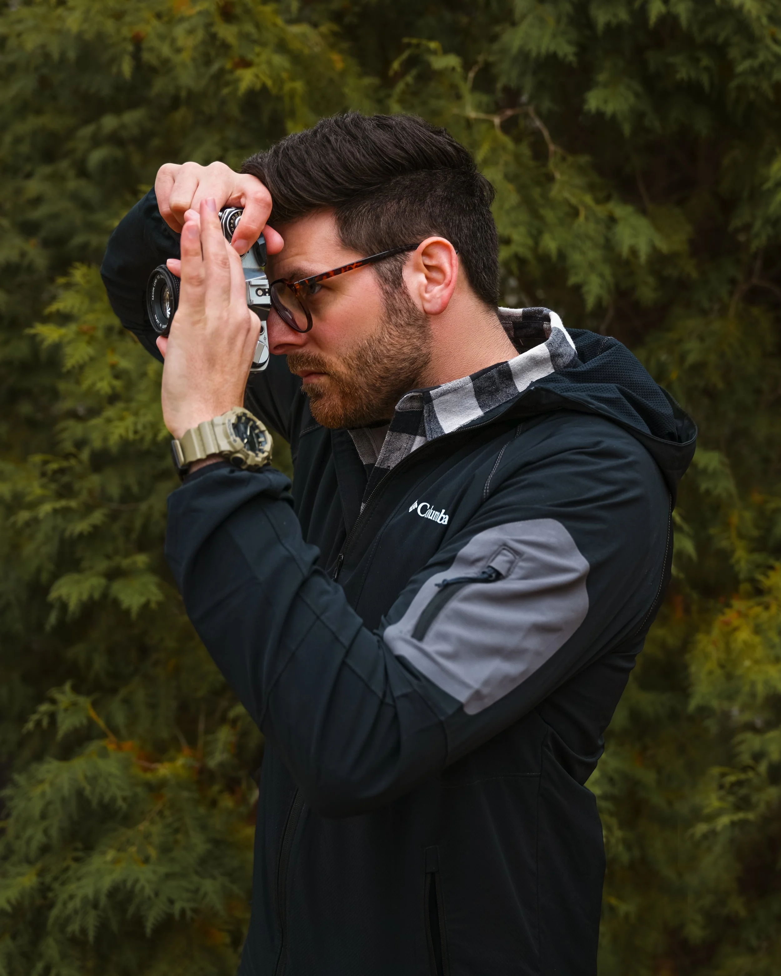 Man with glasses and a beard taking a photograph outdoors with a camera, standing against a backdrop of green trees outside his boudoir photography studio in Calgary..
