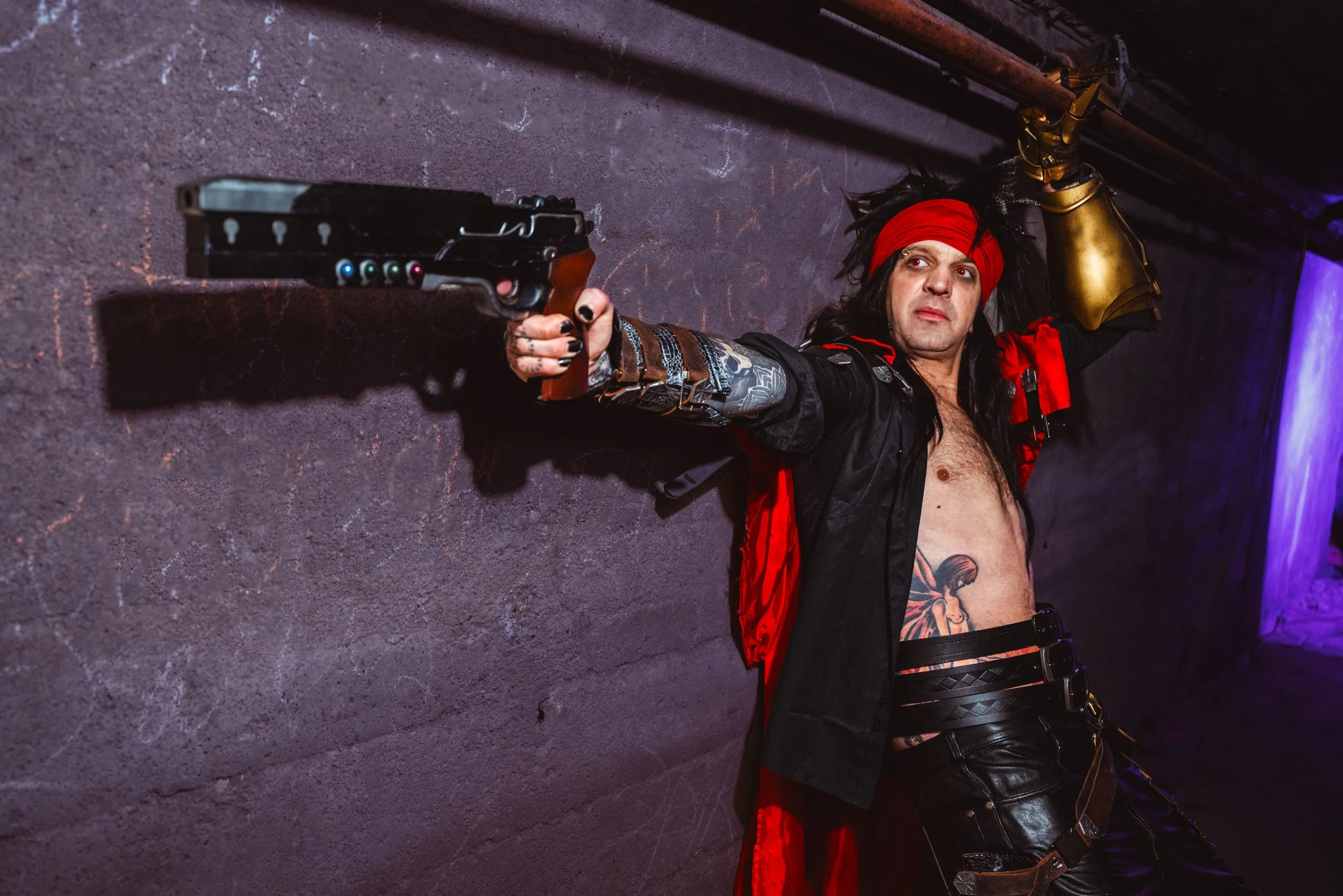 A man dressed in cosplay as a Vincent Valentine from Final Fantasy with long hair, a red headband, and tattoos, aiming a fake gun against a dark wall.