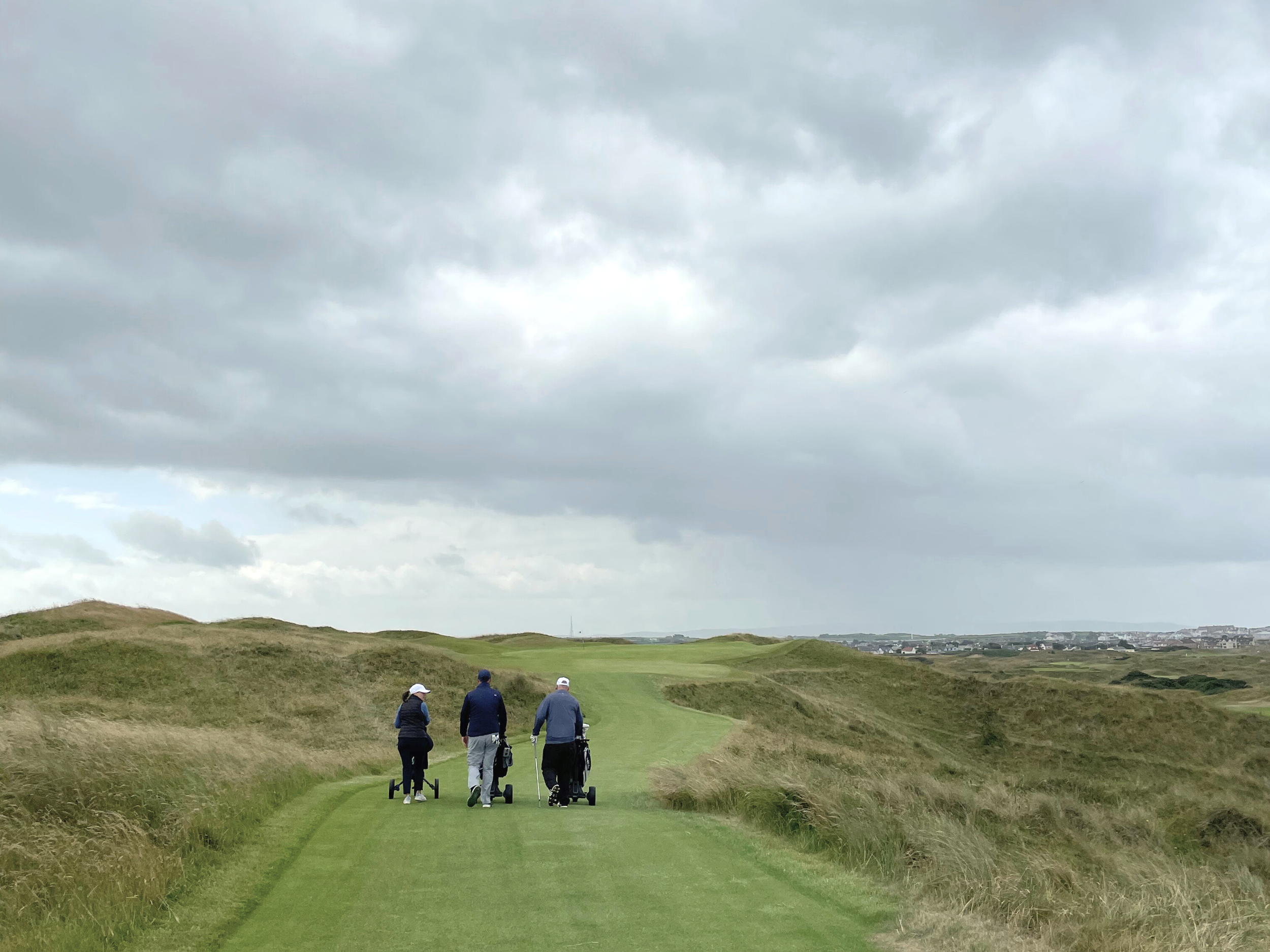 Three people walking on a golf course fairway with golf bags, green grass, rolling hills, and cloudy sky in the background.