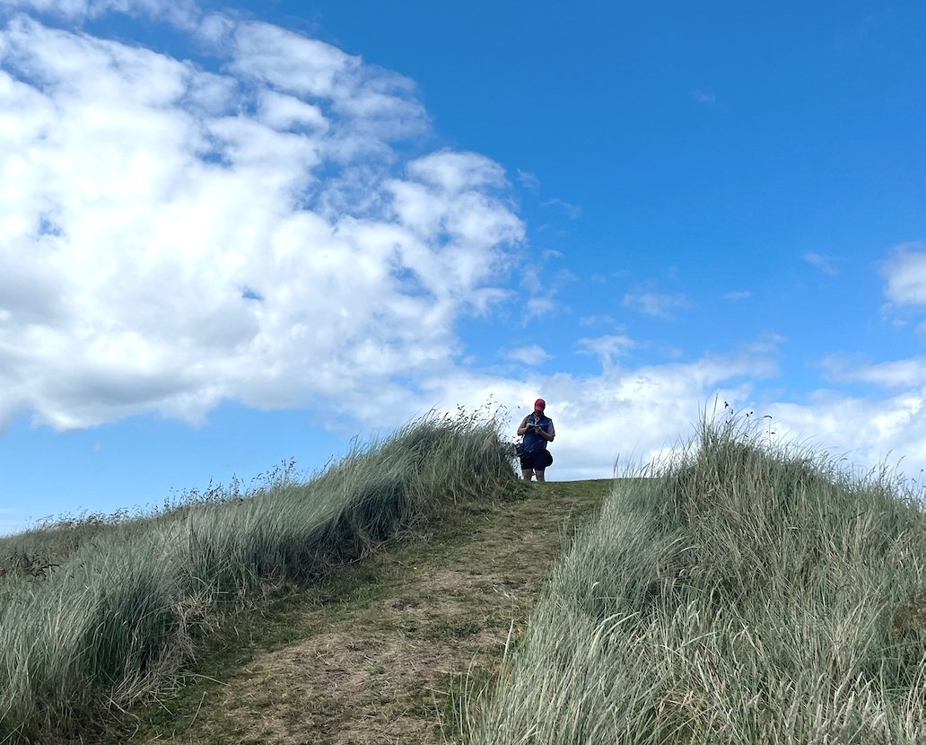 Person walking through grassy dunes under blue sky with clouds