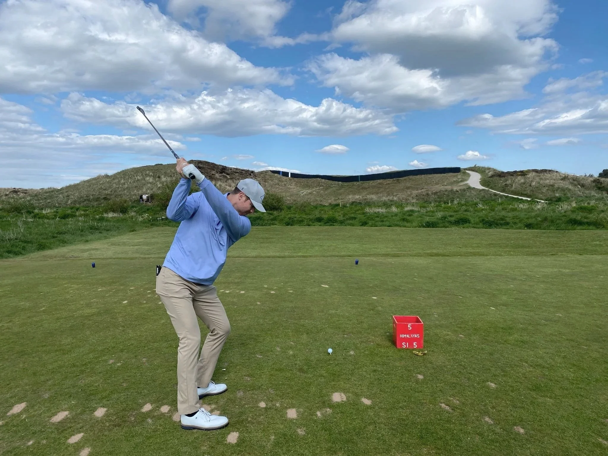 A man playing golf at Prestwick Golf Club, mid-swing with a golf club, under a partly cloudy sky, with rolling hills and a walking path in the background.