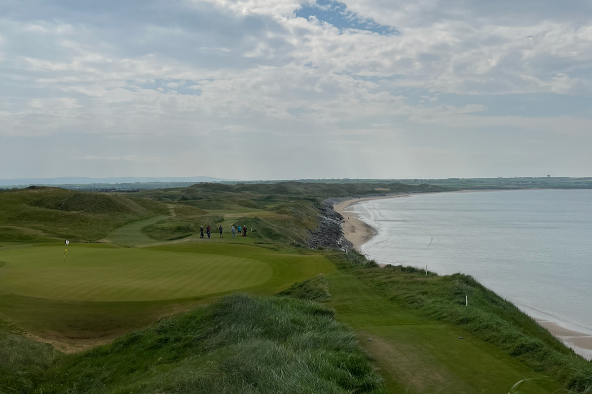 Dark green dunes line the water, with golfers visible in the distance.