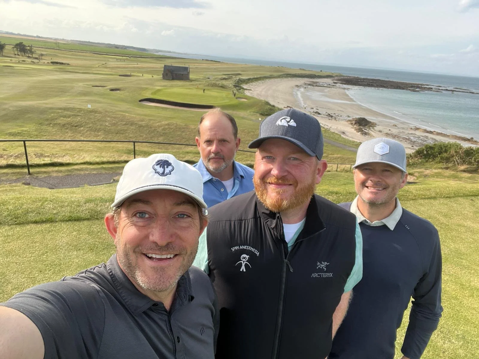 Four men taking a selfie on a golf course with a coastal landscape in the background, featuring grassy fairways, sand traps, and the ocean.