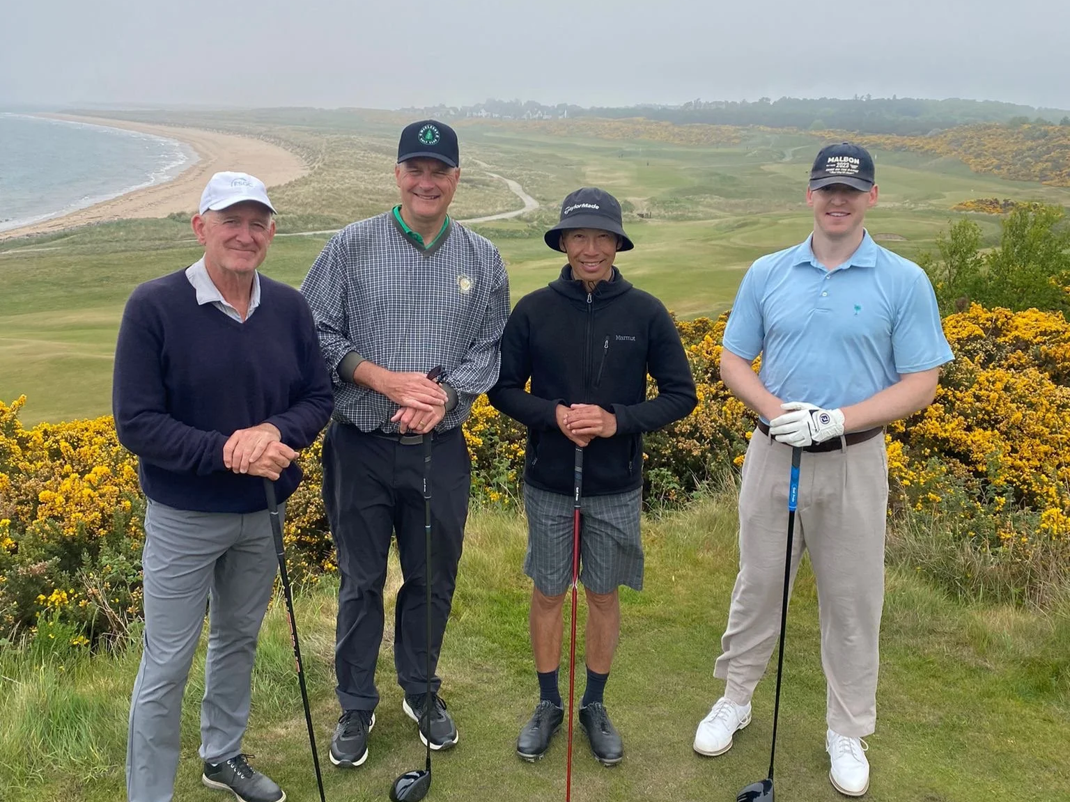 Four men standing on Royal Dornoch Golf Club with yellow blooming bushes behind them, holding golf clubs, smiling, with a beach and ocean in the background.