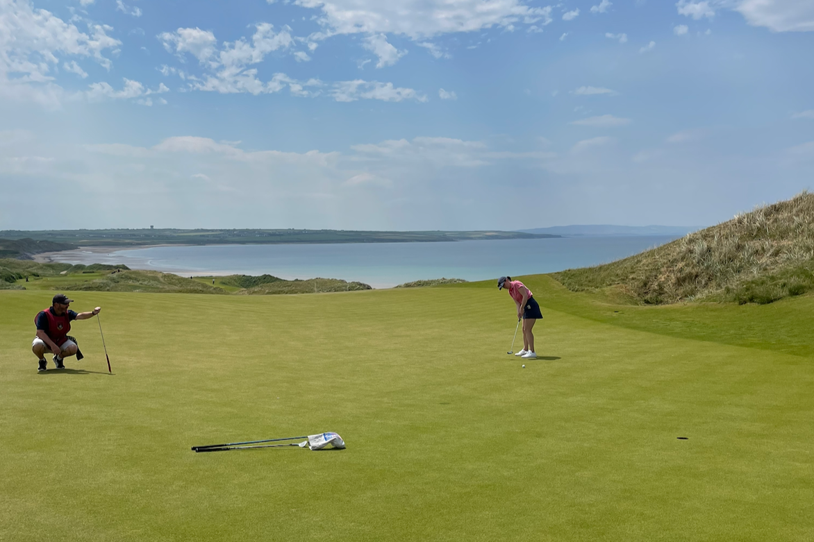 A woman in a pink polo and navy skirt putts on a green at Ballybunion, with blue skies and ocean views in the distance.