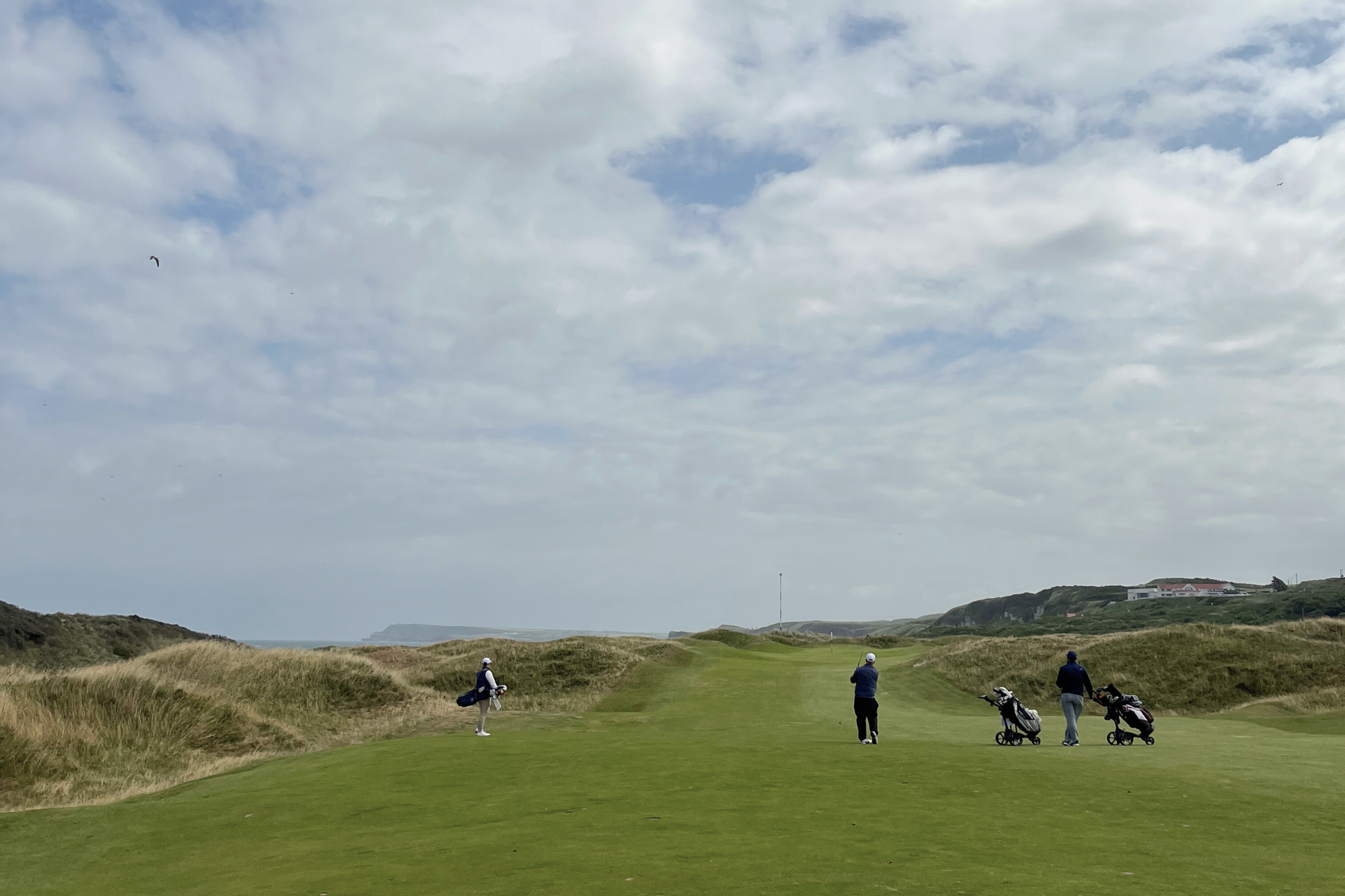 A golfers hits an approach shot into a green on Royal Portrush, with cloudy skies overhead.