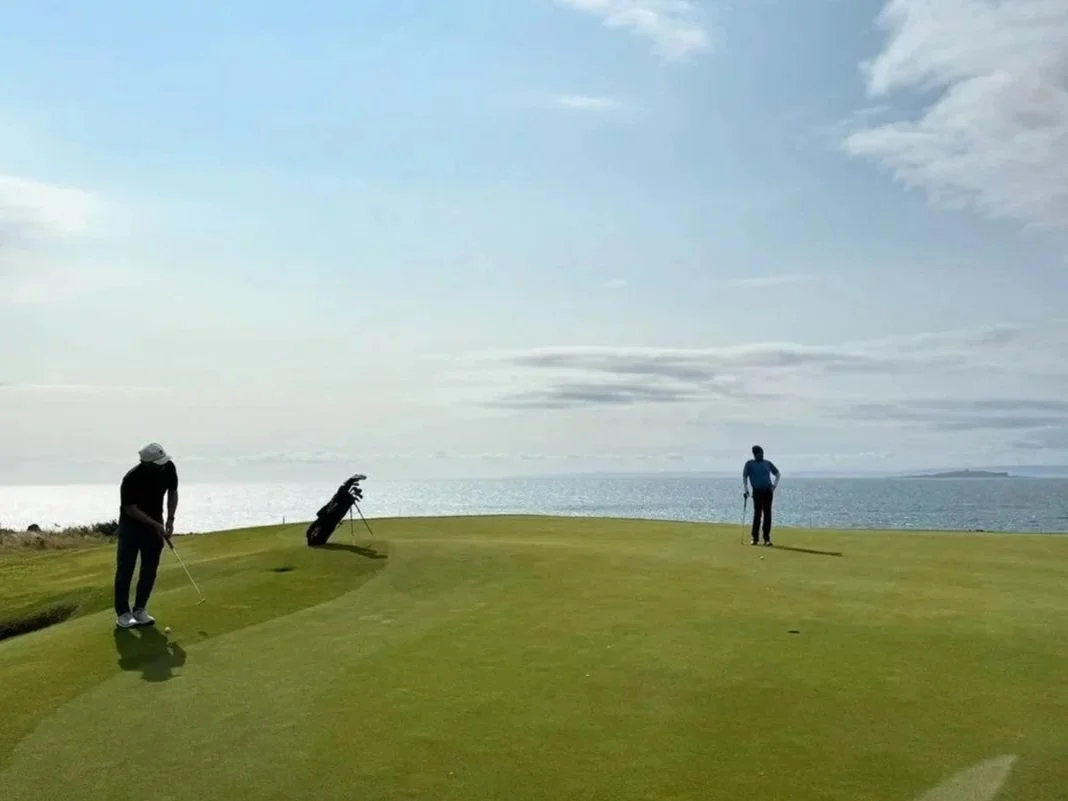 Two men standing near a green overlooking the ocean in the background.