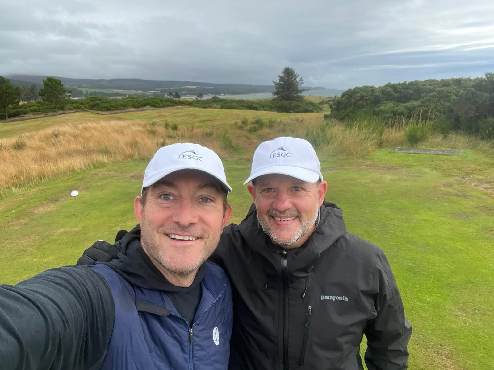 Two men smiling and taking a selfie on a golf course, wearing white ESGC hats, with a scenic landscape and cloudy sky in the background.