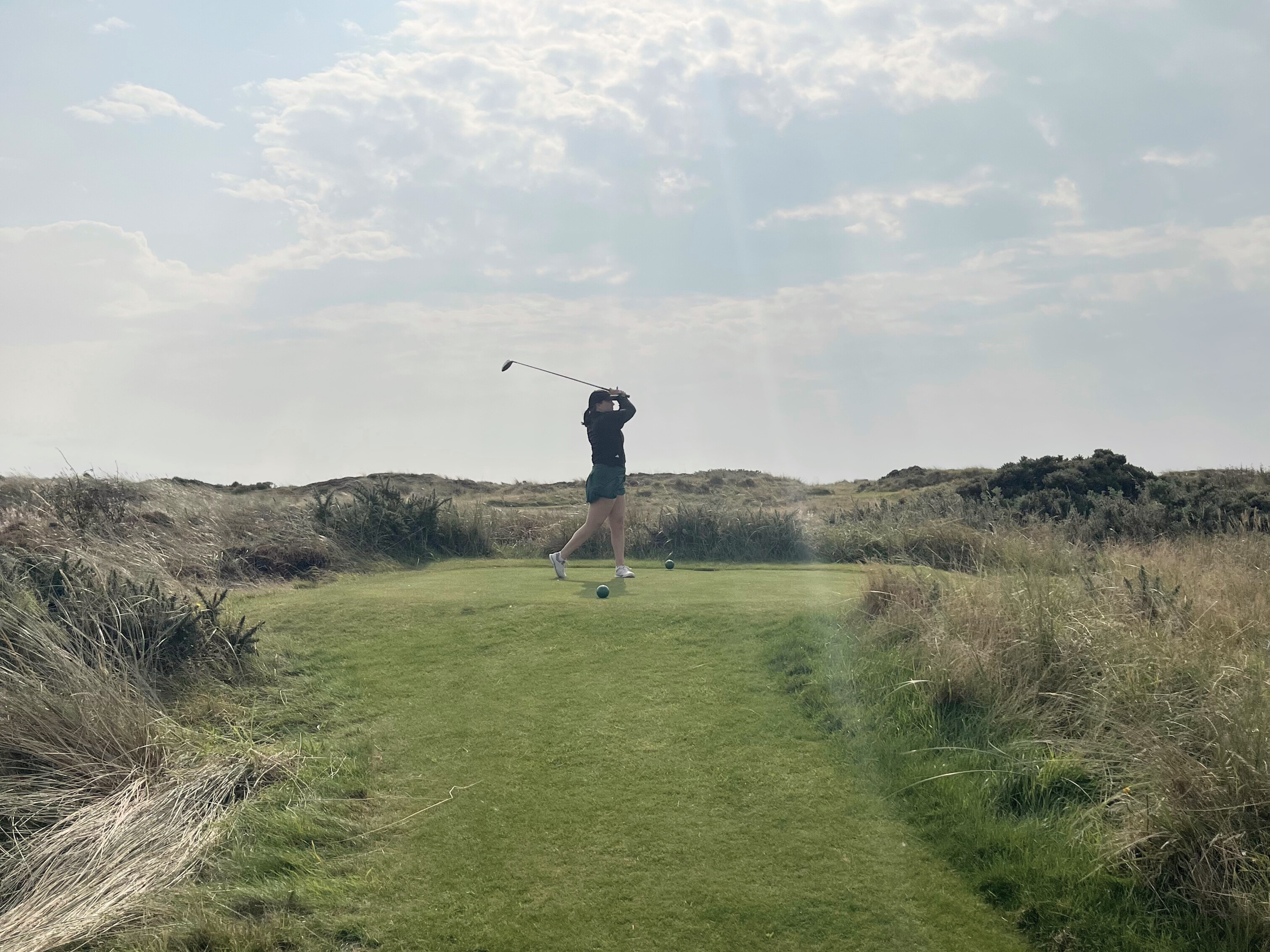 A person standing on a tee at Western Gailes Golf Club preparing to hit a golf ball on a golf course surrounded by grassy dunes and under a partly cloudy sky.