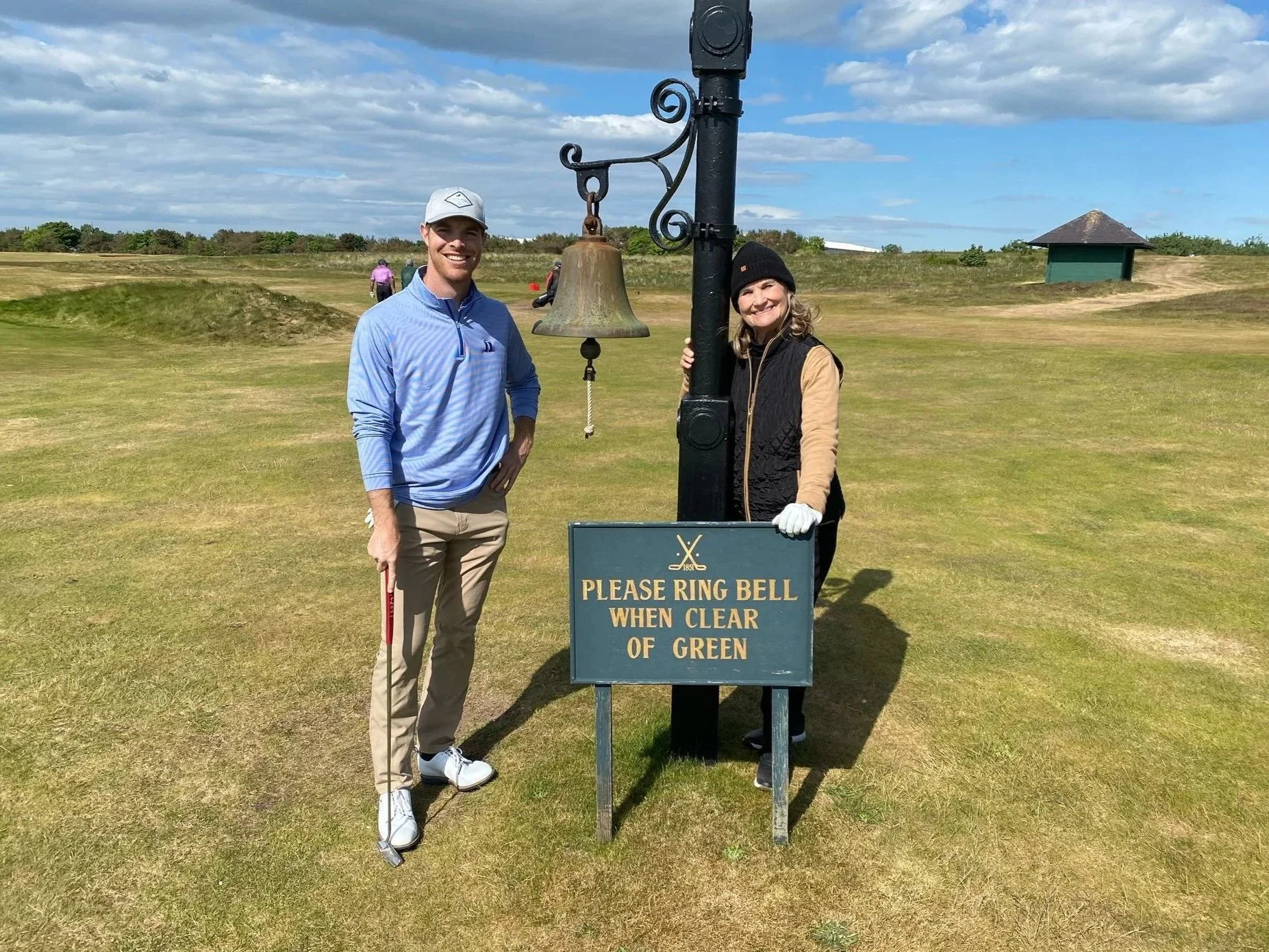 Two people standing next to a bell on a golf course. The man is holding a golf club, smiling, wearing a gray cap and a blue striped long-sleeve shirt. The woman is wearing a black beanie, a beige long sleeve shirt, and white gloves, smiling and holding the bell's rope. There is a sign in front that says, 'Please ring bell when clear of green.' The background features a grassy golf course, a small green building, and a partly cloudy sky.