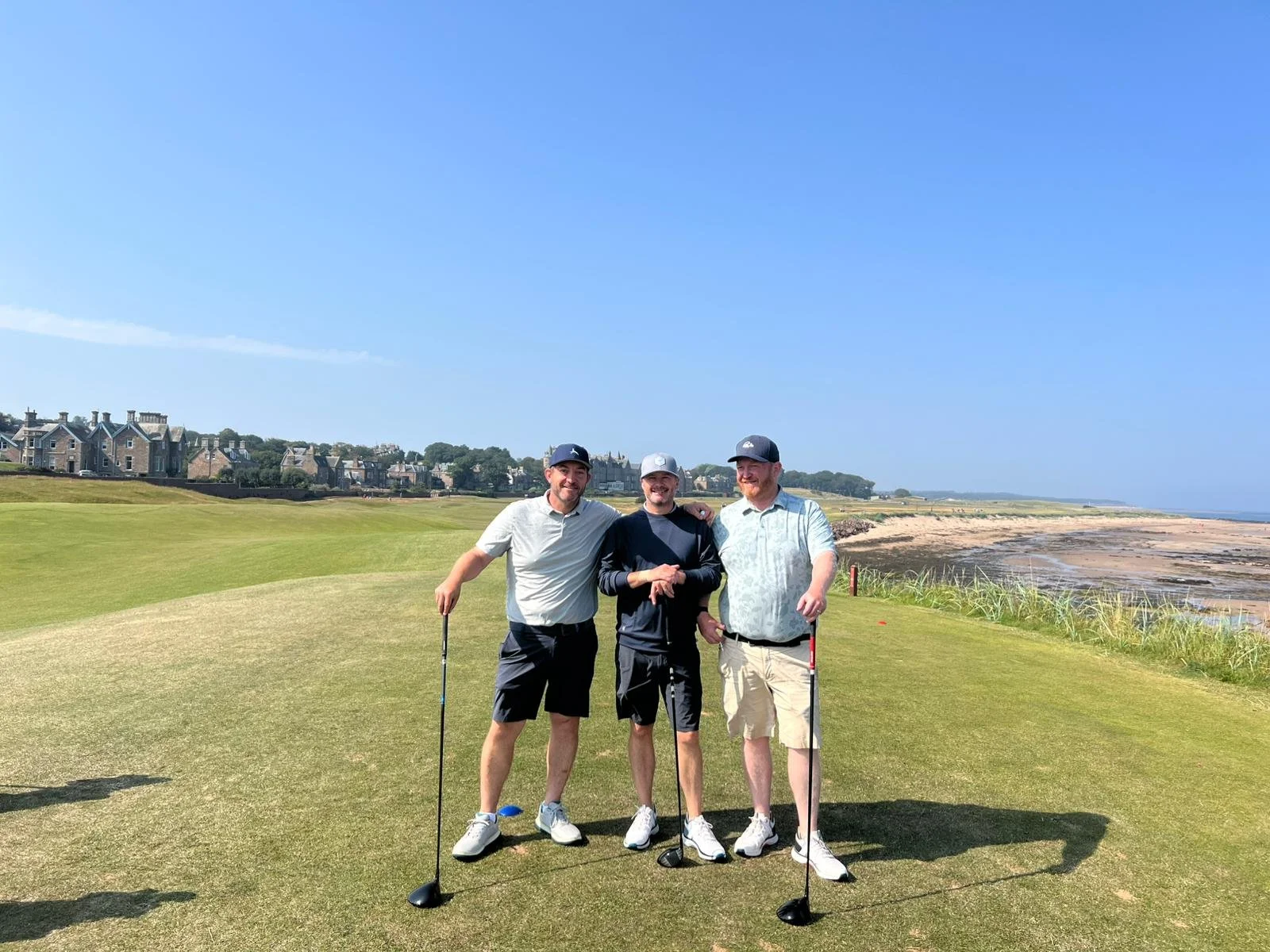 Three men standing on a golf course near the coast, holding golf clubs, smiling at the camera, under clear blue sky.