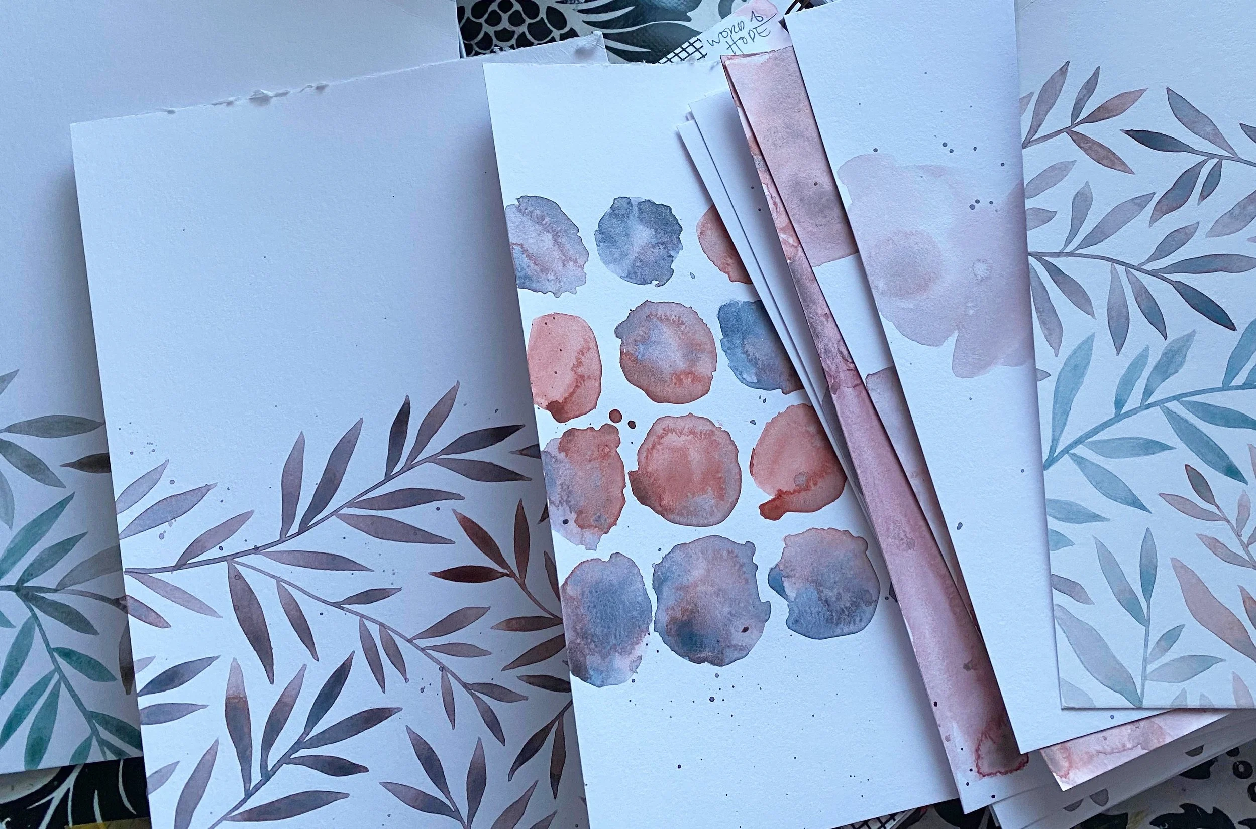Assorted watercolor painted paper sheets with botanical and abstract designs, layered on top of each other.
