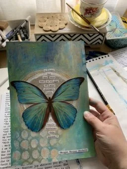 A hand holding an open journal or book with a large depiction of a blue butterfly on the cover. The background features abstract, colorful patterns with blue and green hues, and there are various art supplies and objects on a desk nearby.