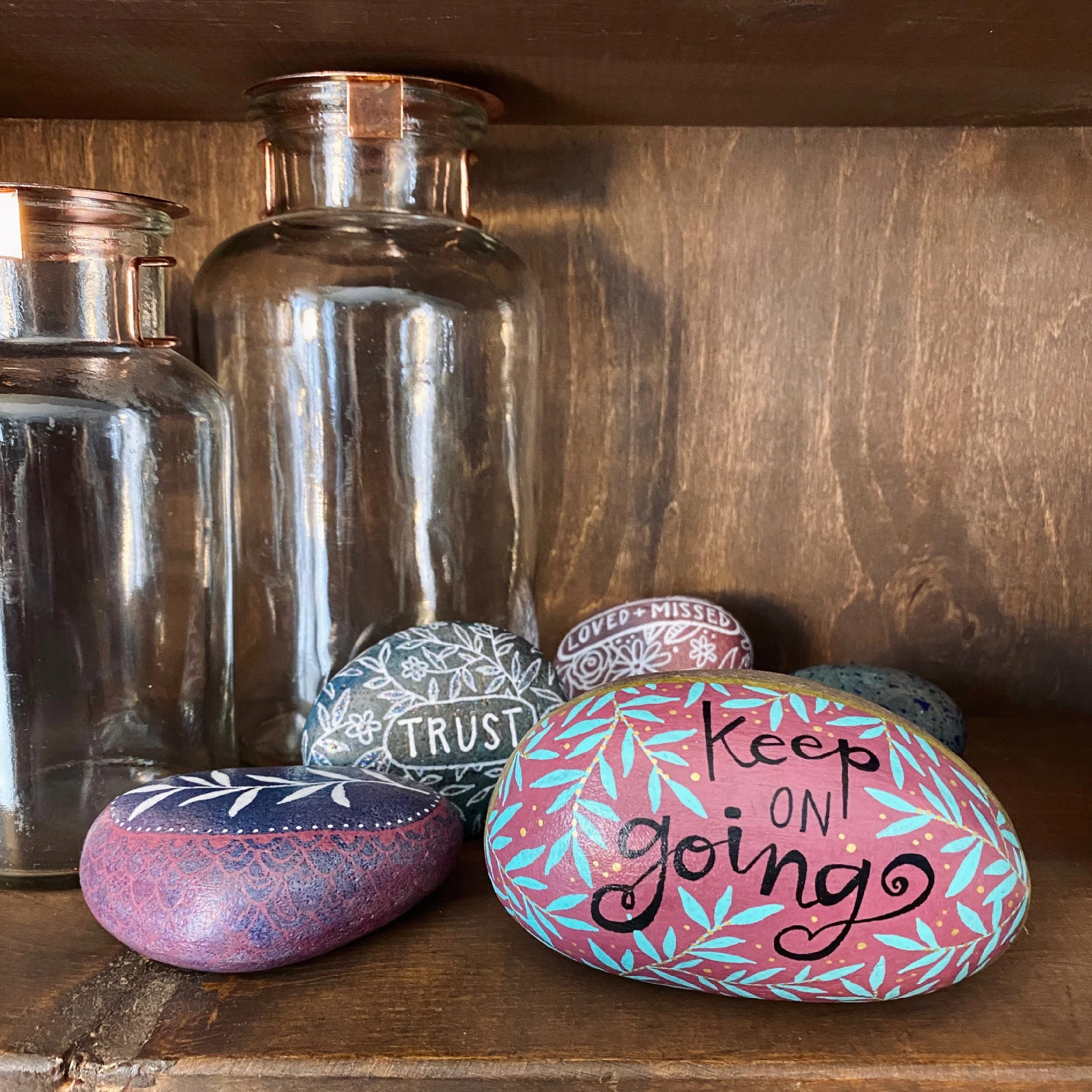 Colorfully painted rocks with inspirational messages such as 'Keep on going,' 'Trust,' and 'Loved + Missed,' arranged on a wooden shelf alongside glass containers.