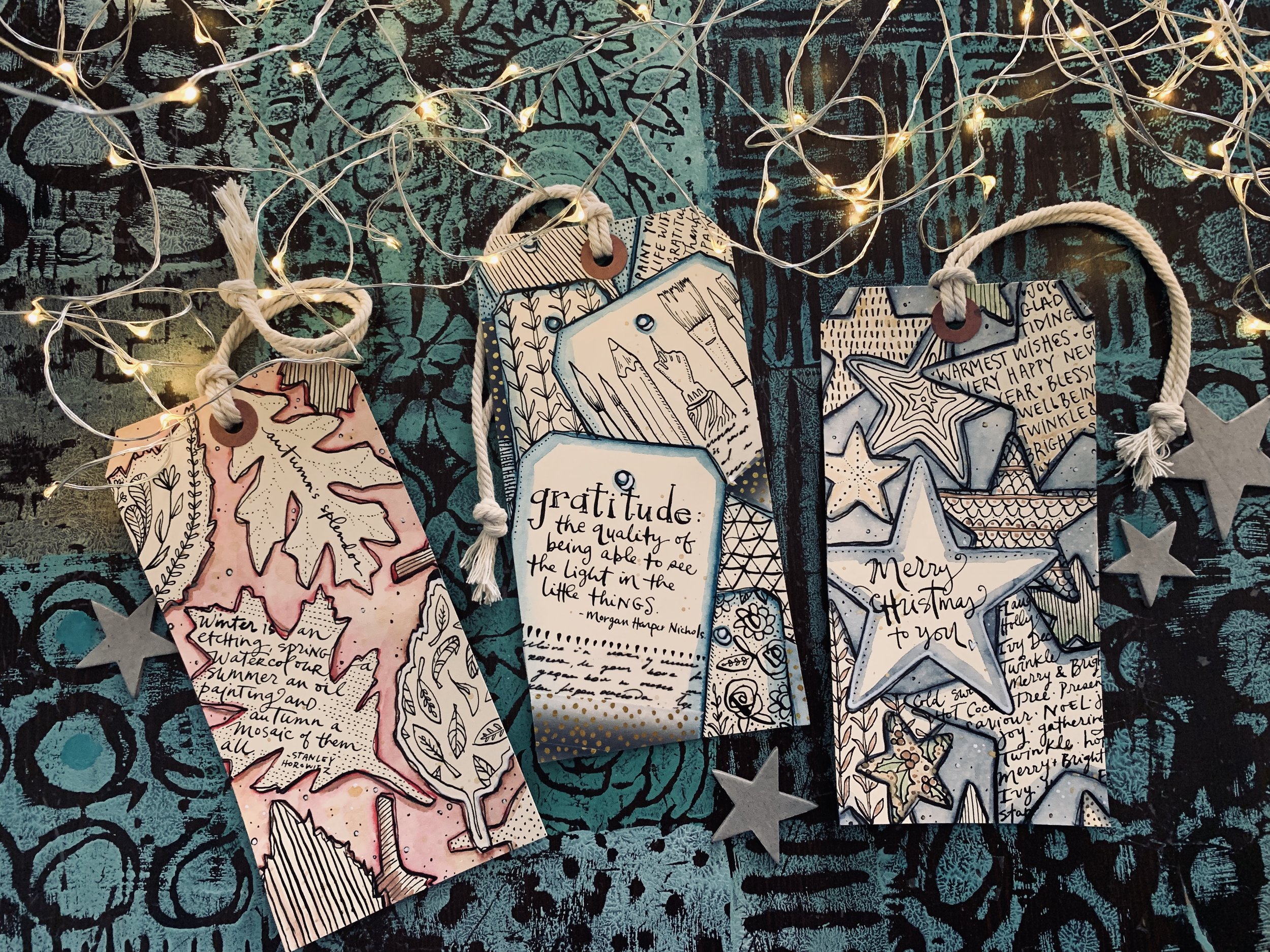 Set of decorative holiday tags with handwritten messages, illustrations of stars, trees, leaves, and a quote about gratitude, placed on a textured dark background with string lights.