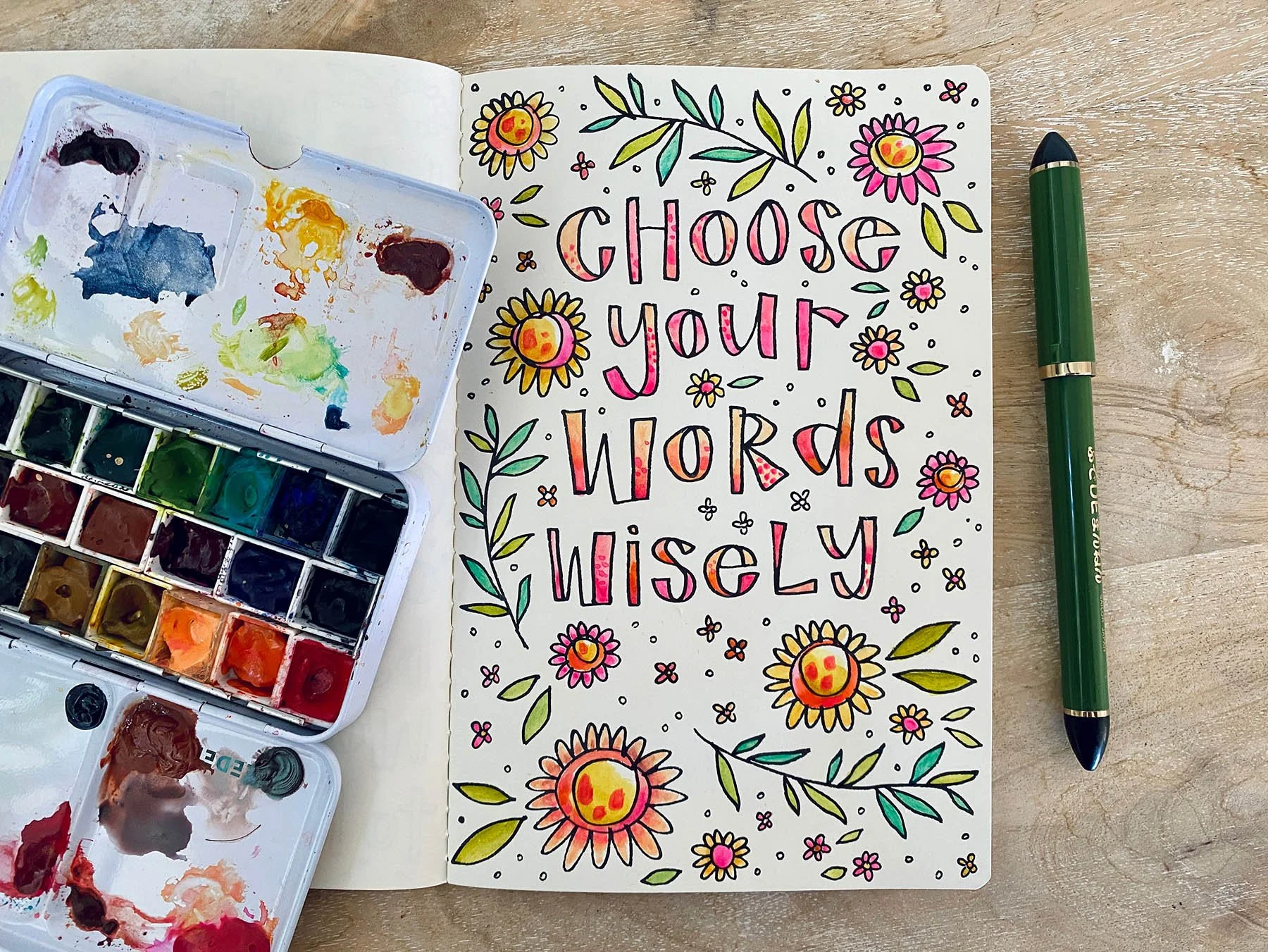 Colorful hand-lettered sign with the words "Choose Your Words Wisely" surrounded by flowers and decorative elements, next to watercolor paints and a green pen on a wooden surface.